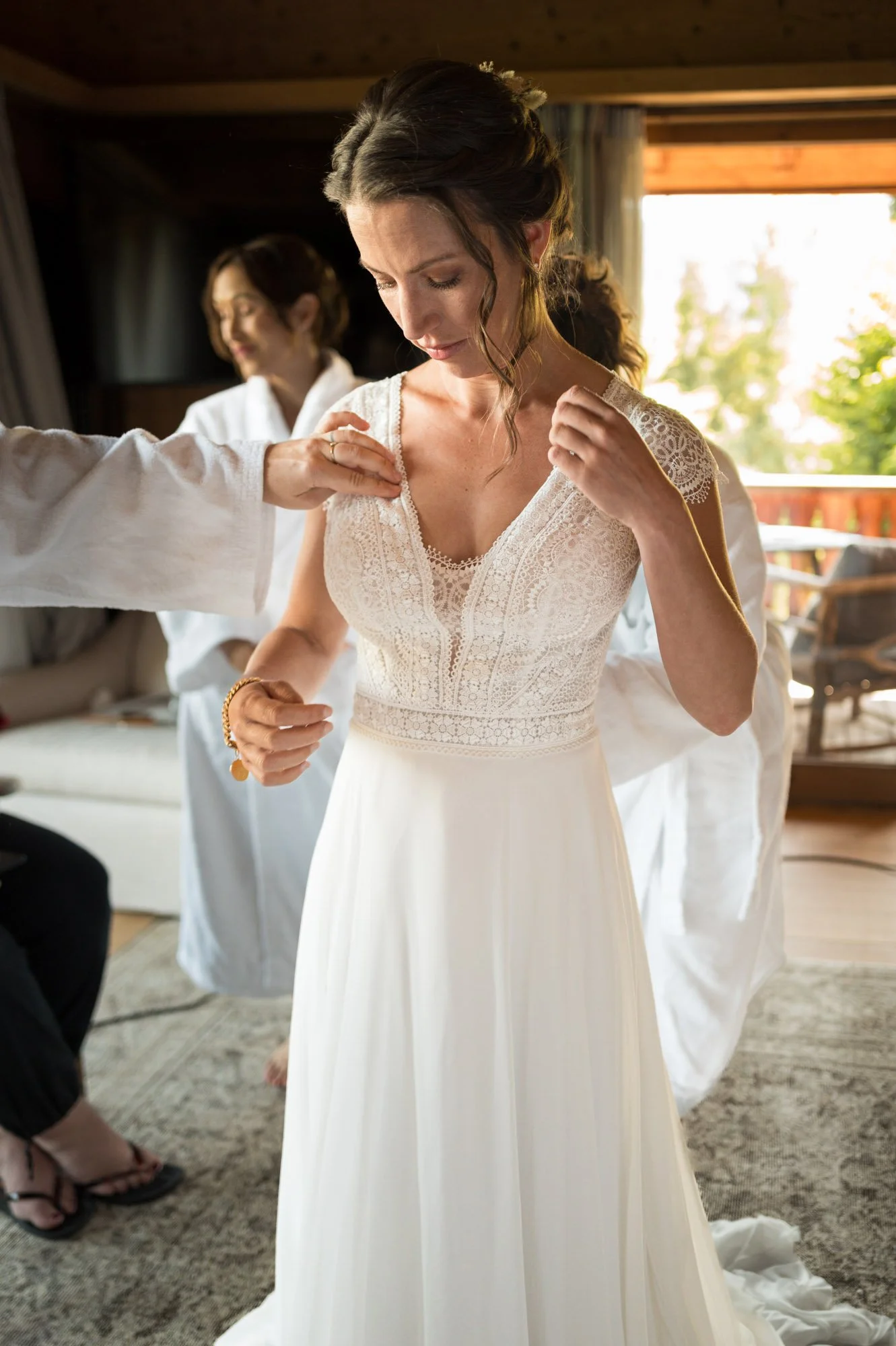 Fée de mariage en train d'essayer une robe de mariée, avec deux autres femmes en arrière-plan, dans une pièce lumineuse avec vue sur l'extérieur.