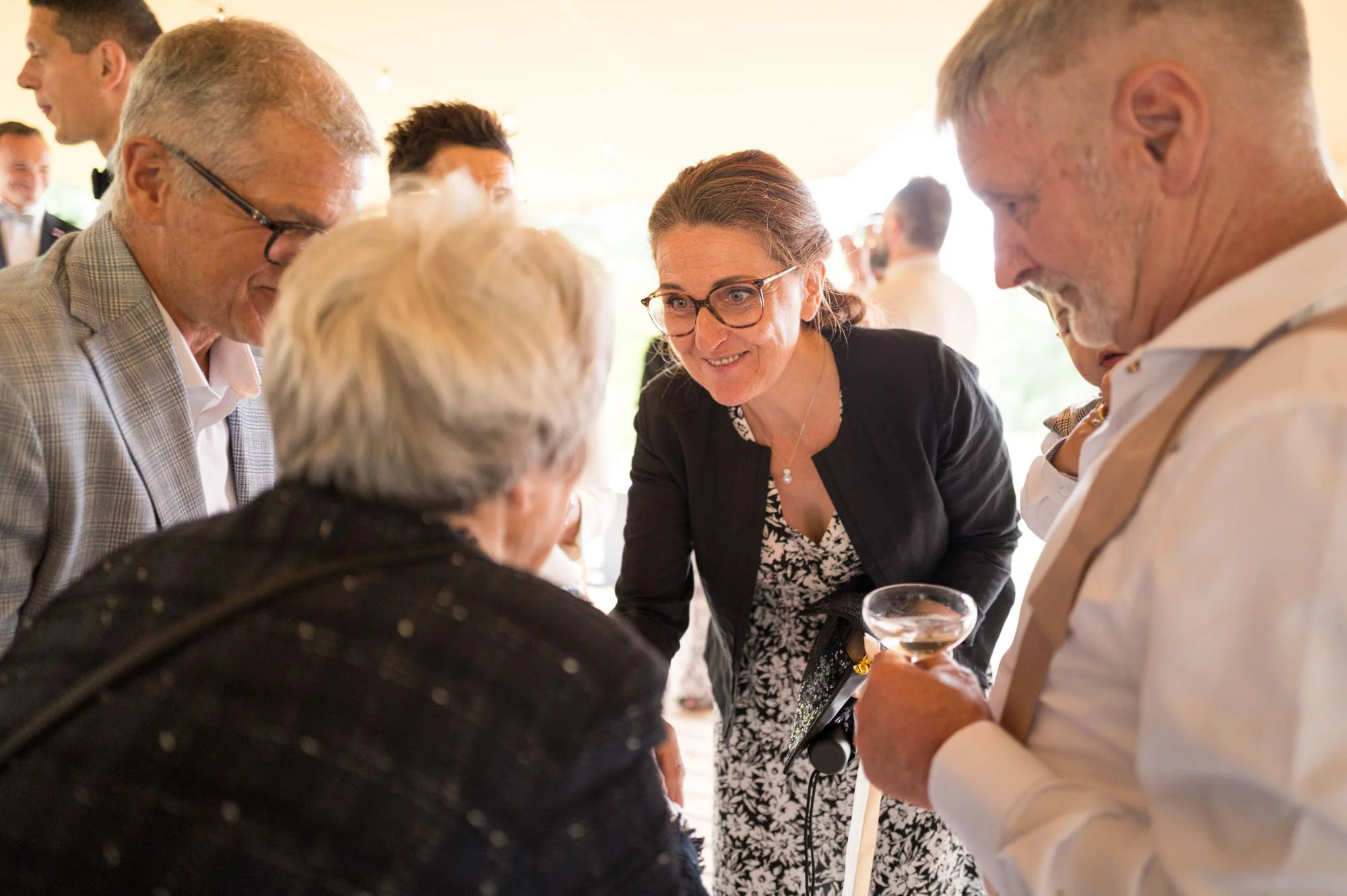 Un groupe de personnes en conversation lors d'une célébration, avec un homme avec un verre à la main, un homme avec des lunettes, un femme avec des lunettes et une amie plus âgée