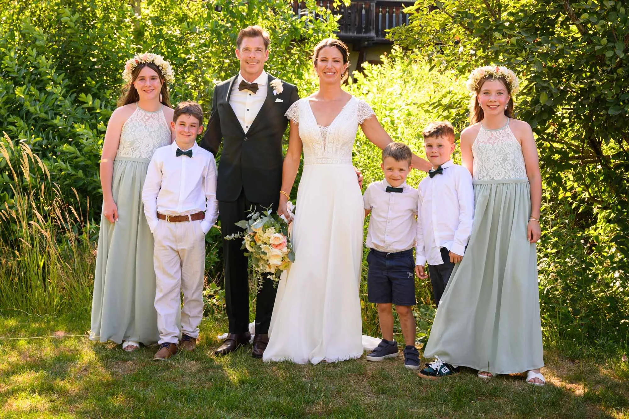 un groupe de personnes posant pour une photo lors d'un mariage en plein air, comprenant un couple marié, trois enfants et deux jeunes filles portant des robes longues avec des couronnes de fleurs.