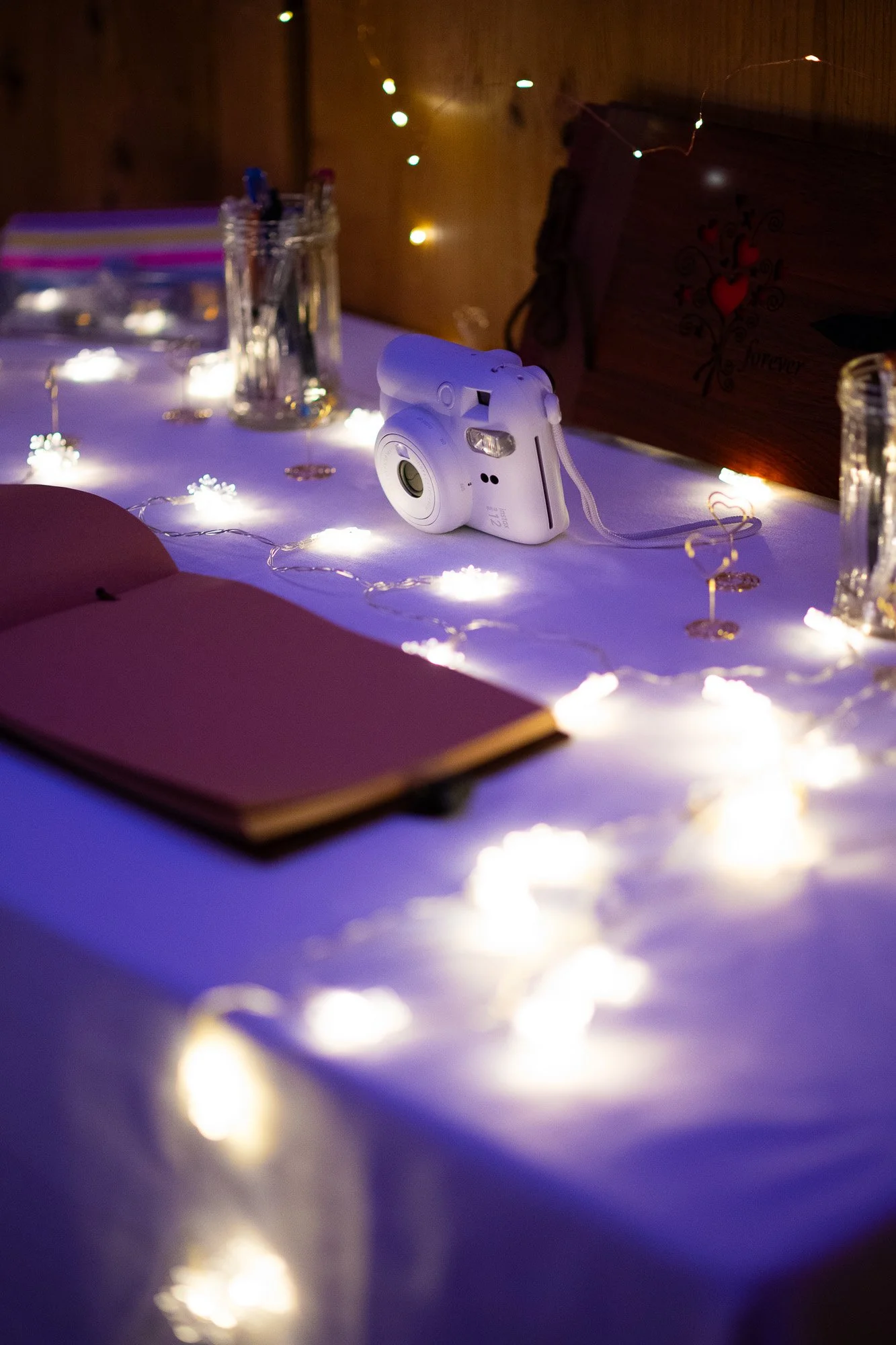 Une table décorée avec des guirlandes lumineuses blanches, une caméra blanche, un cahier rose, des verres en verre, et une boîte en bois avec un motif de cœur rouge.