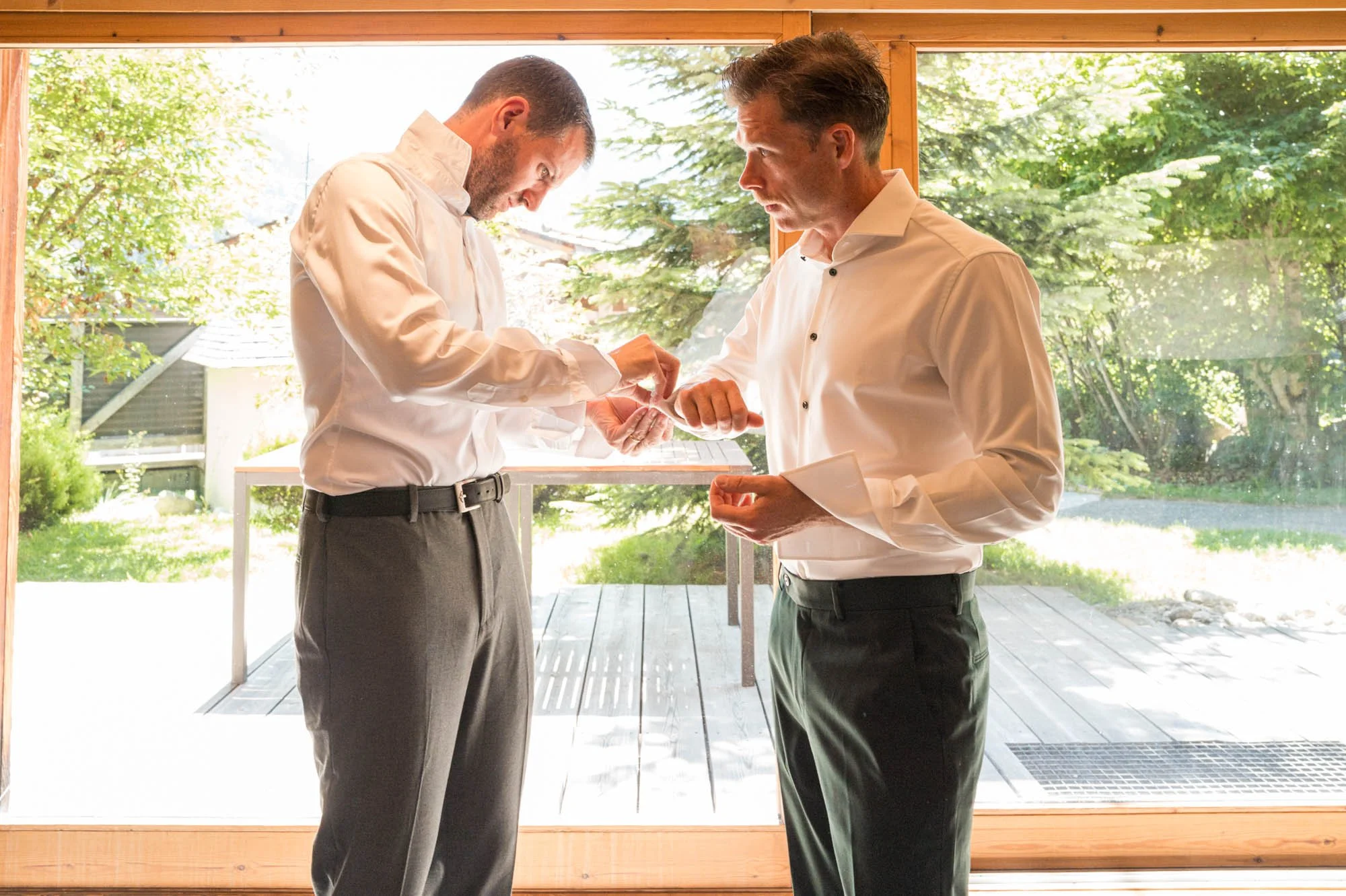 Deux hommes en chemises blanches et pantalons noirs discutent devant une grande fenêtre donnant sur un jardin vert