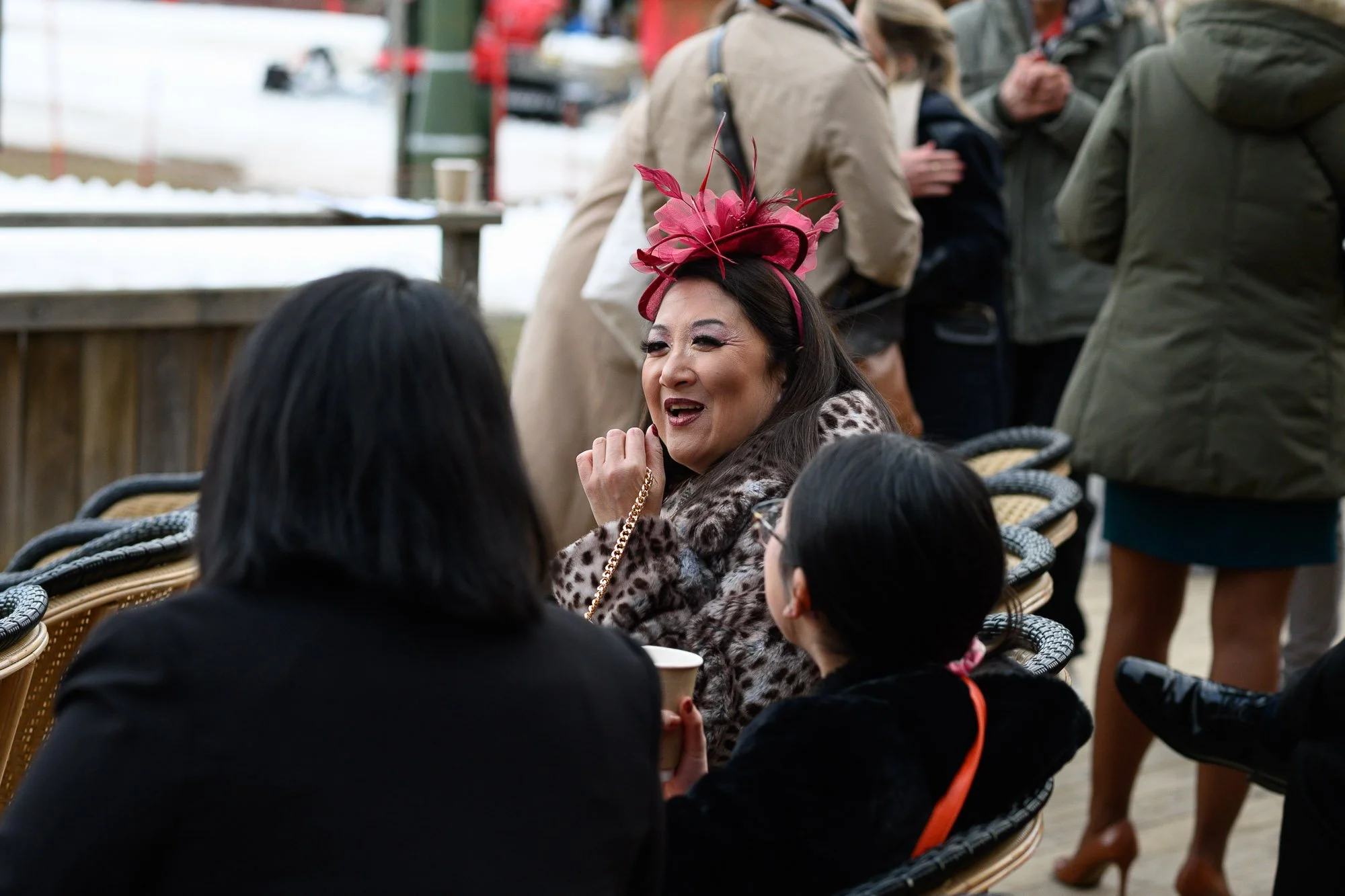 Une femme souriante portant un chapeau rouge en plumes discute avec deux autres personnes assises à une table lors d'une fête en plein air, avec des gens qui se tiennent en arrière-plan.