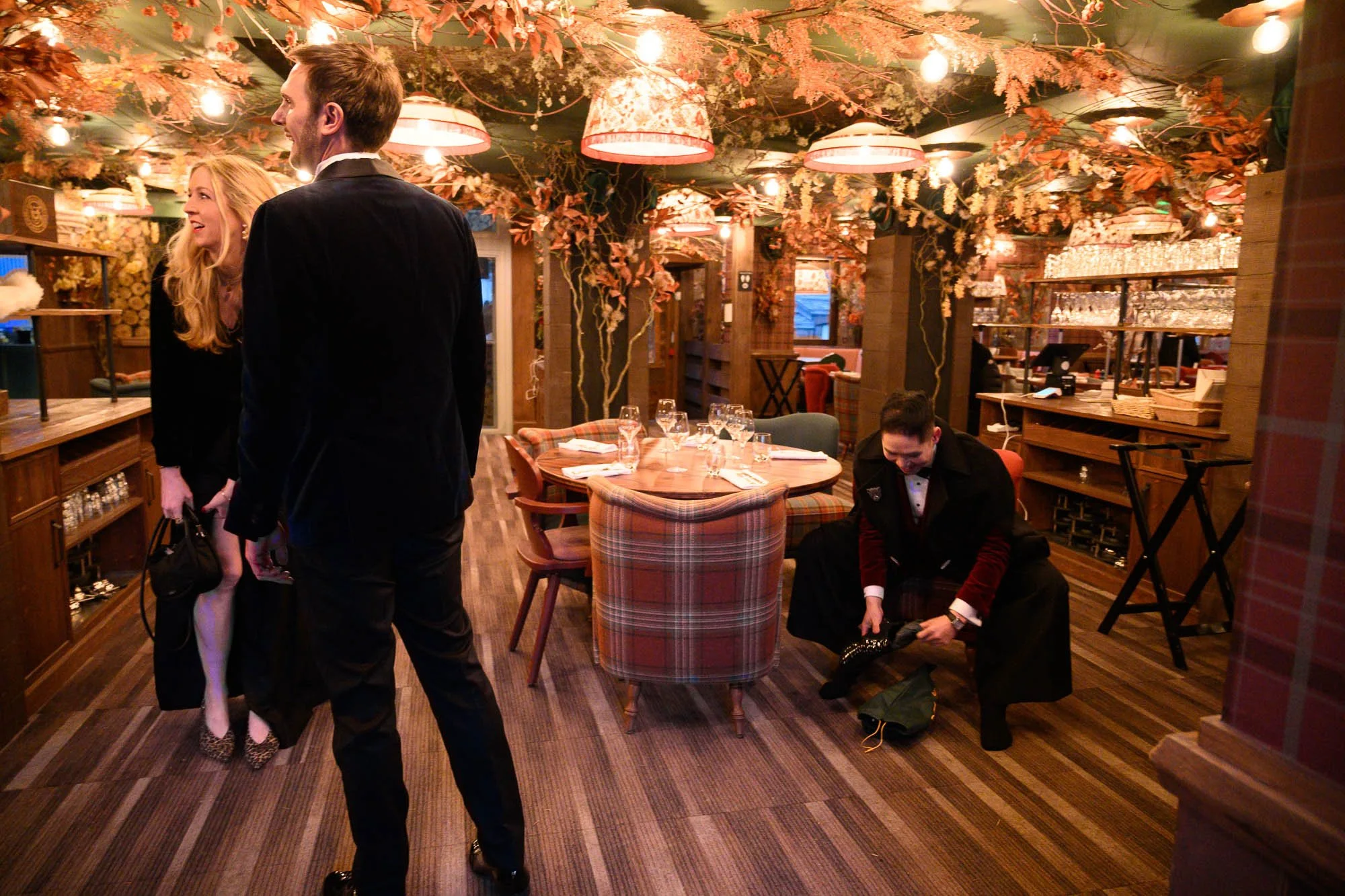 Une scène dans un restaurant chic avec des décorations automnales, où un homme et une femme élégamment habillés s'engagent dans une conversation, tandis qu'un serveur ajuste ses chaussures à côté d'une table. L'ambiance est chaleureuse et festive ave