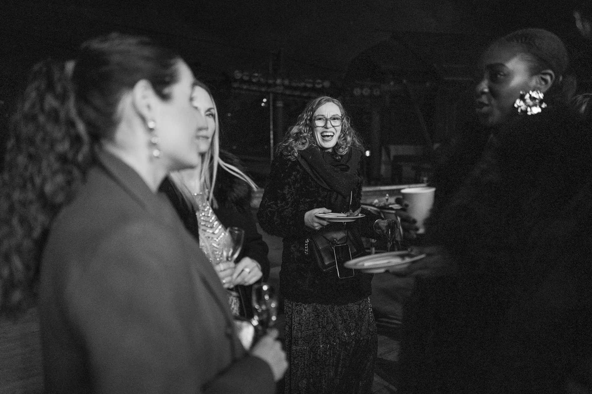 Un groupe de femmes en conversation, au centre une femme avec lunettes sourit et parle, autres femmes écoutent. Ambiance de soirée ou événement social.