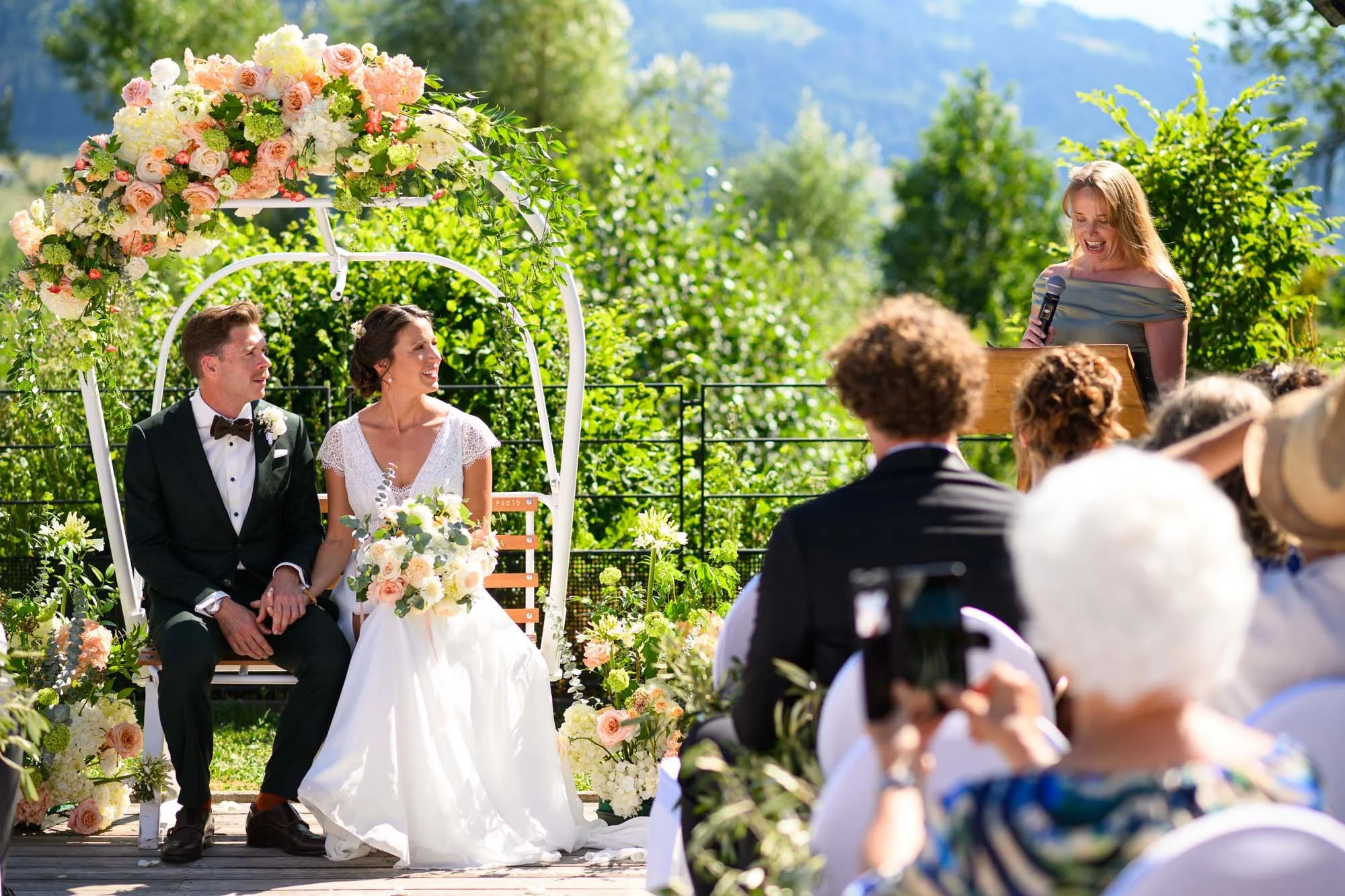 Cérémonie de mariage en plein air avec un couple assis sous une arche fleurie, femme en robe blanche et homme en costume sombre, devant un groupe d'invités, avec une officière parlant dans un micro, en pleine nature