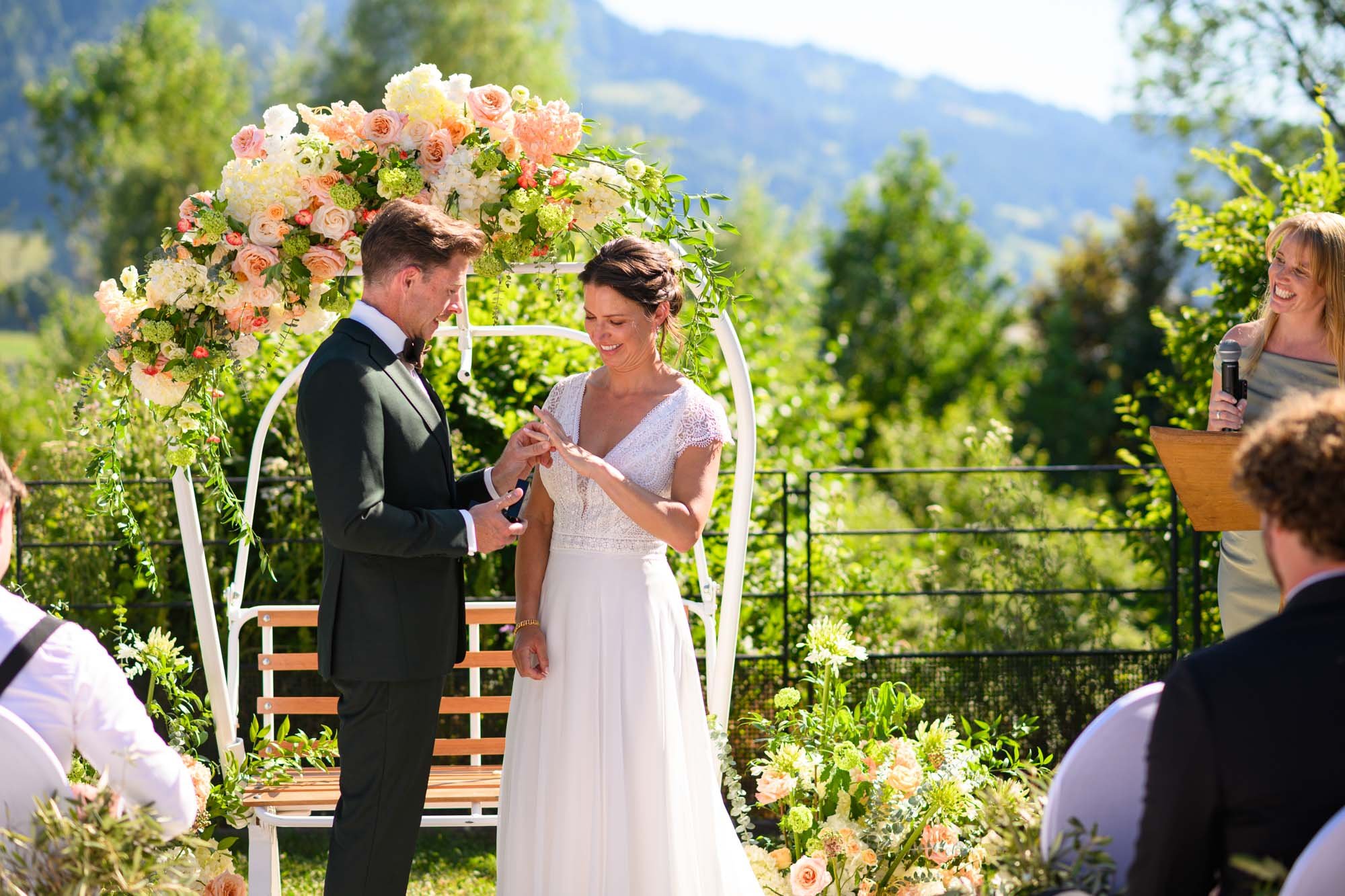 Un couple en tenue de mariage échange des anneaux lors d'une cérémonie en plein air sous un ciel ensoleillé, entouré de fleurs et de verdure, avec une officiante souriante tenant un micro.