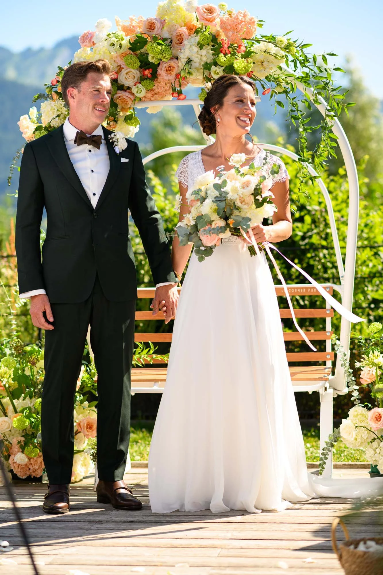Un couple de mariés en tenue de mariage se tenant la main lors d'une cérémonie en plein air, entouré de fleurs et sous un canopy floral.