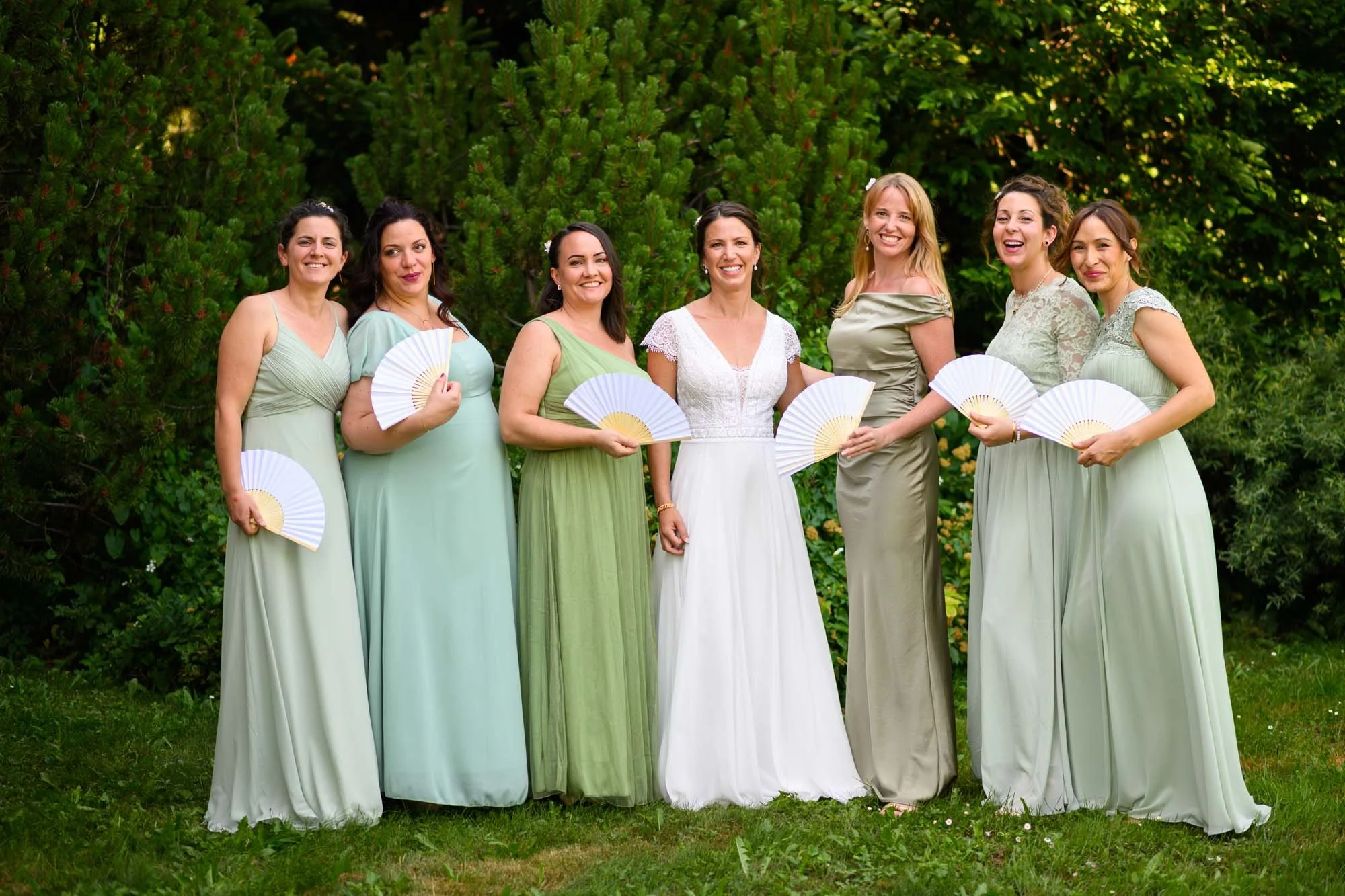 Groupe de femmes en robes de mariée vert clair ou blanche, tenant des éventails blancs, posant devant des arbustes verts lors d'une cérémonie de mariage en plein air.