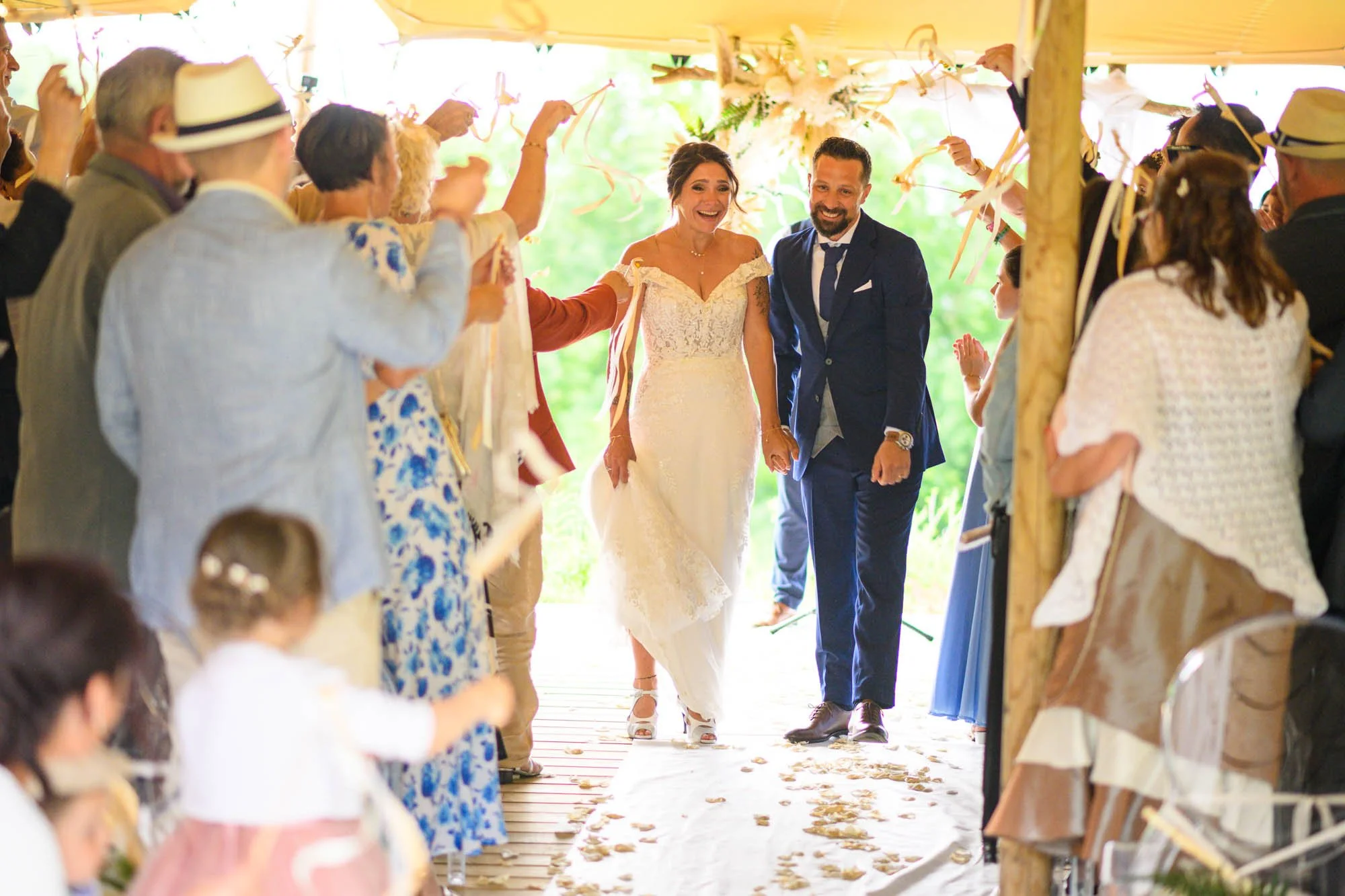 Un couple marié marche main dans la main lors de leur mariage sous une tente décorée, entouré d'invités qui les acclament et agite des rubans.