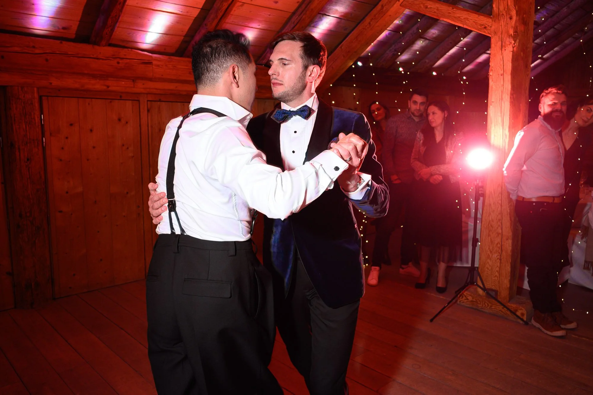 Deux hommes dansent lors d'une fête. L'un porte une chemise blanche avec des bretelles noires, l'autre un costume noir avec une veste en velours bleu et un nœud papillon. En arrière-plan, d'autres personnes regardent la danse, dans un décor en bois a