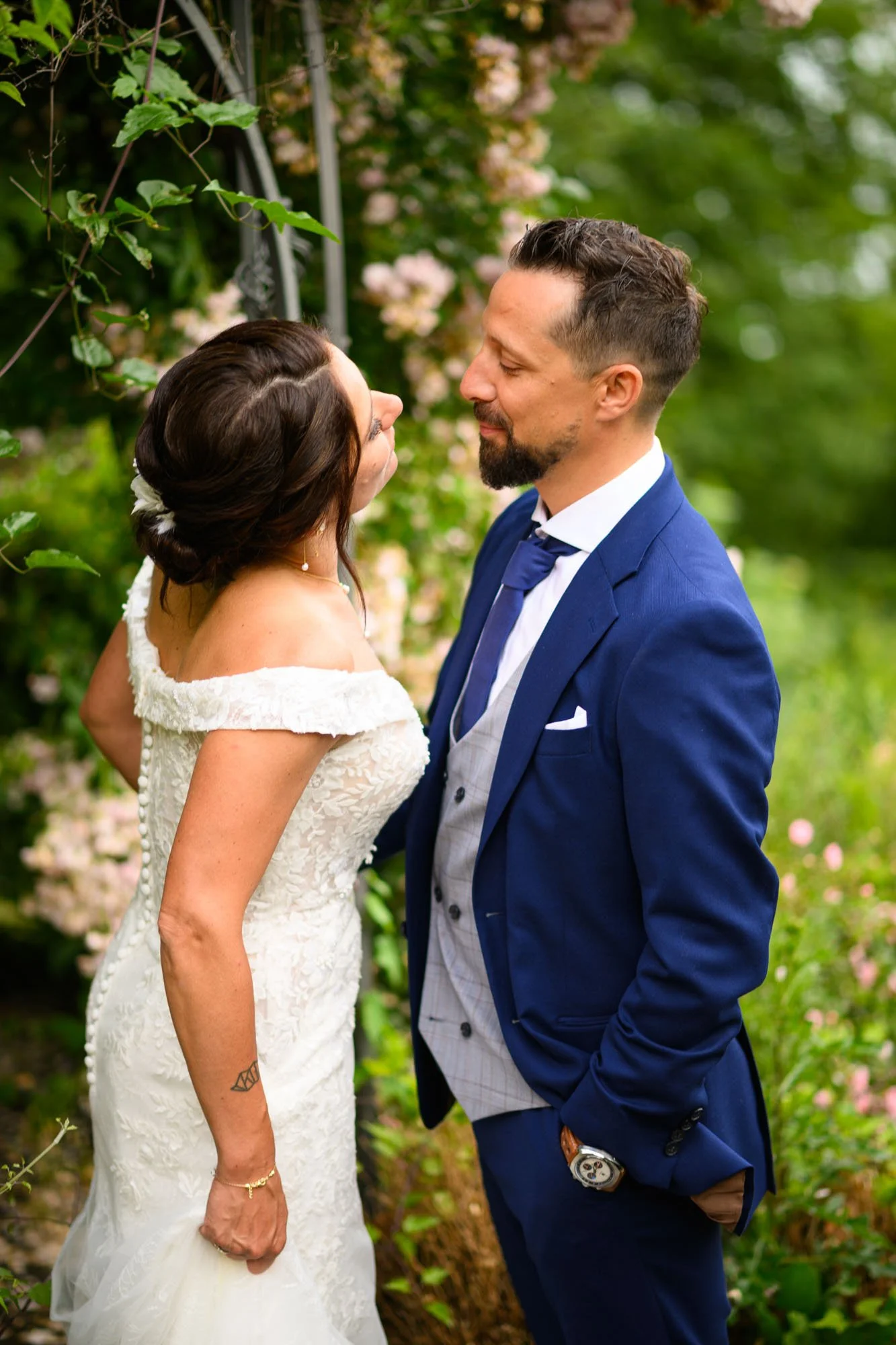 Un couple de mariés se regarde dans un jardin verdoyant avec des fleurs roses, souriants et proches, la femme en robe blanche et le homme en costume bleu.