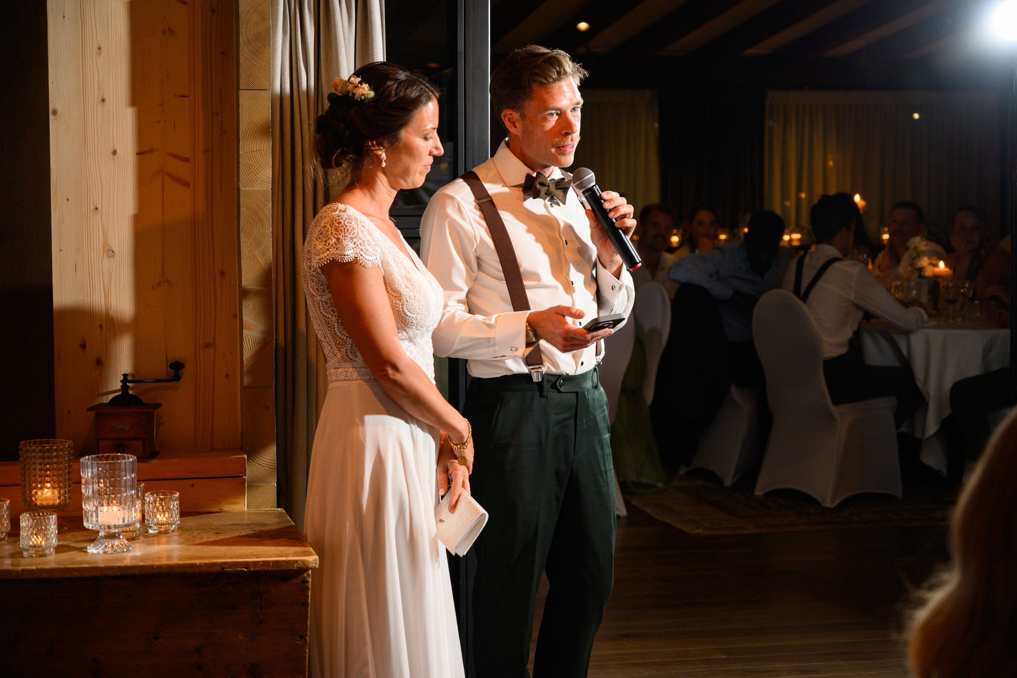 Un couple lors d'un discours de mariage, la femme porte une robe blanche en dentelle et le homme porte une chemise blanche, un pantalon foncé avec des bretelles, ils se tiennent à côté d'une table en bois avec des bougies. En arrière-plan, des invité