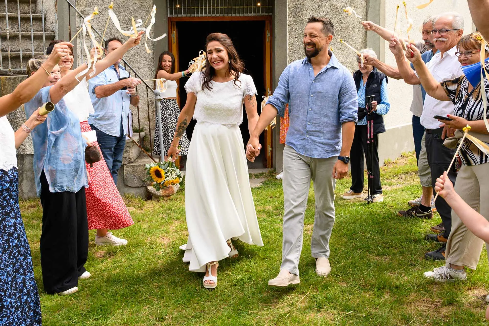 Un couple de mariés, la femme en robe blanche et le mari en chemise bleue, marche main dans la main à l'extérieur. Ils sont entourés par un groupe de personnes qui leur font une haie d'honneur avec des confettis et des guirlandes, célébrant leur mari