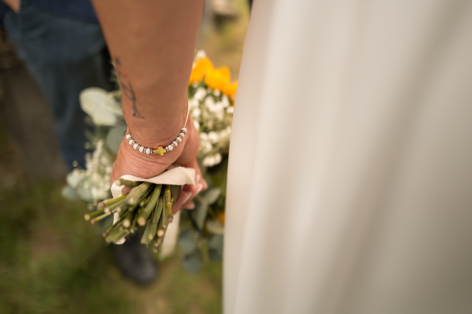 Première personne tenant un bouquet de fleurs, portant un bracelet en perles avec une croix. La photo est prise de dos, lors d'une cérémonie ou un mariage.