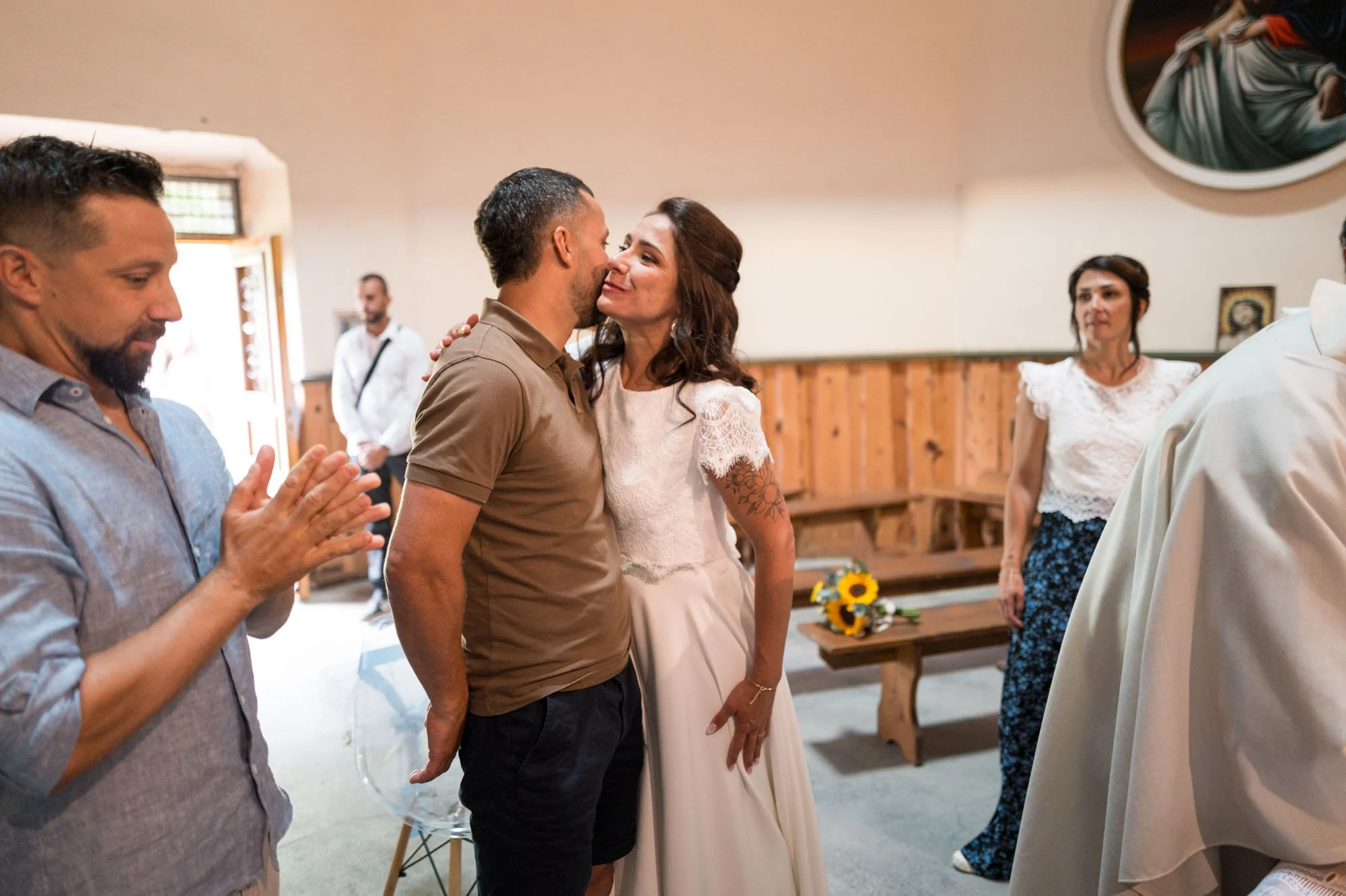 Un couple lors d'une cérémonie, probablement un mariage, s'embrasse tendrement. Sur le fond, quelques personnes observant l'événement.