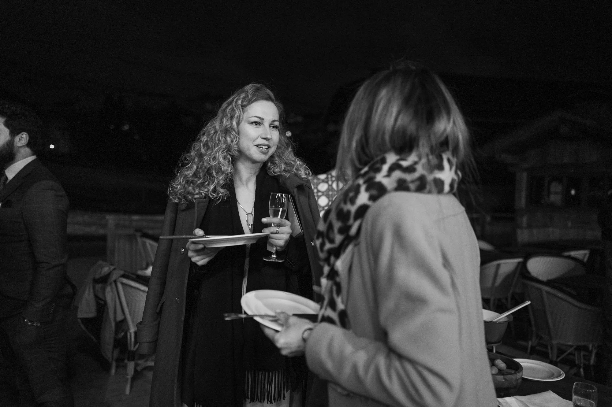 Deux femmes en conversation lors d'une réception, une tenant une flûte de champagne et un plateau, l'autre tenant une assiette. En arrière-plan, un homme en costume et un espace de réception avec des chaises.