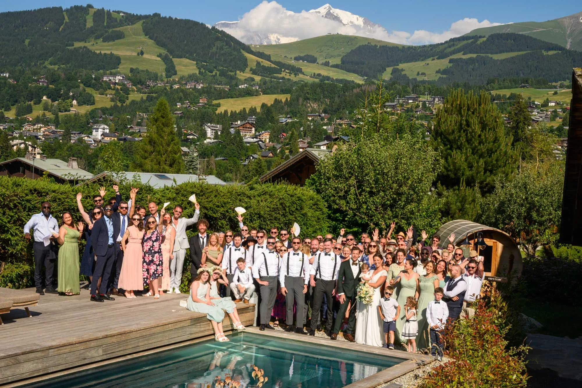 Un groupe de personnes déguisées en costumes pour un mariage, posant pour une photo devant une piscine, avec un paysage montagneux en arrière-plan—labels et accessoires de fête