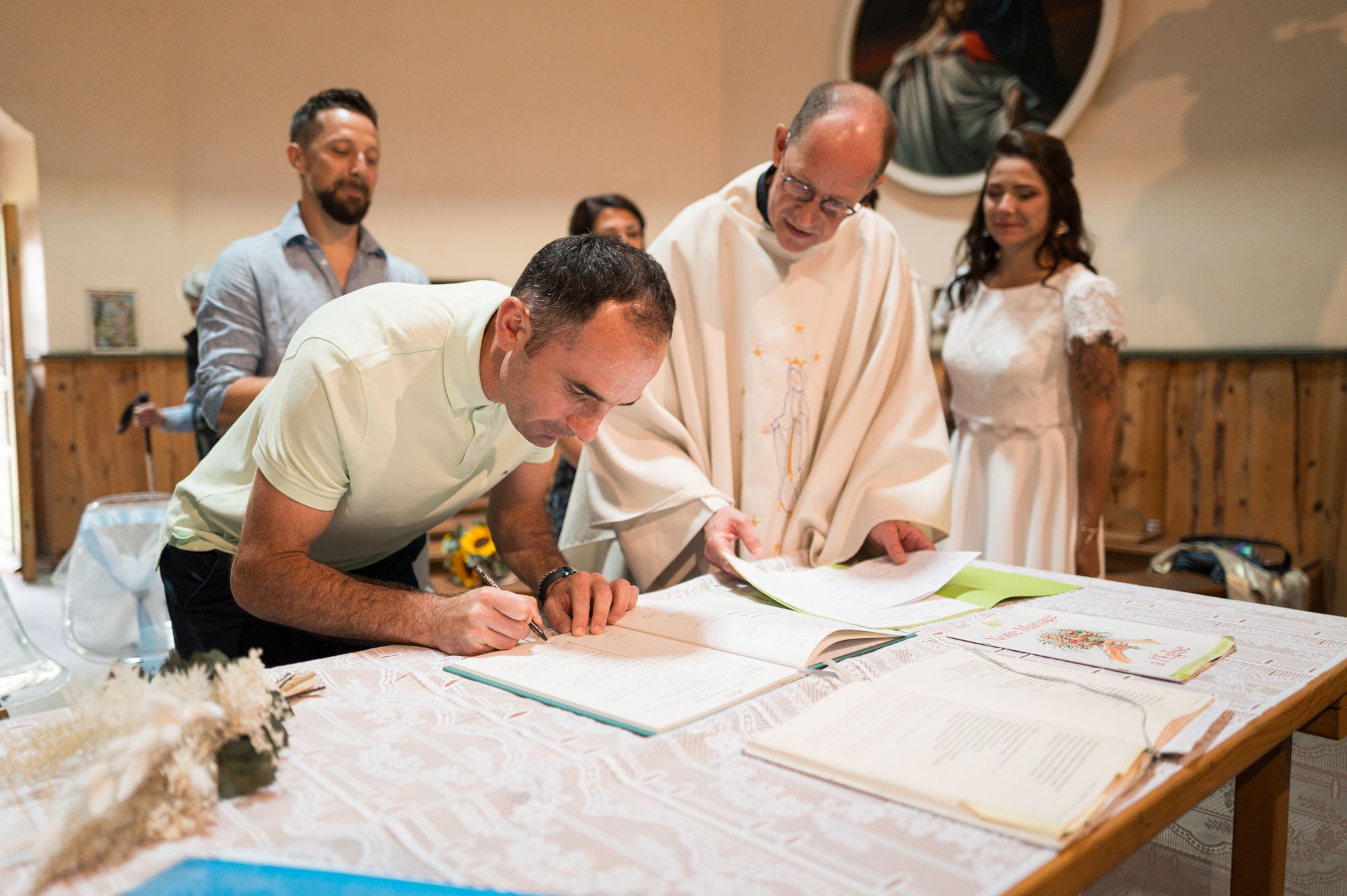 Un homme signe un livre sur une table lors d'une cérémonie religieuse avec un prêtre et d'autres personnes observant, dans un espace intérieur avec décor en bois.