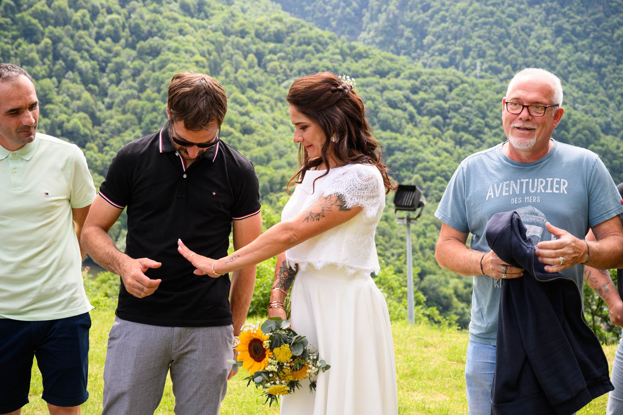 Un groupe de personnes en extérieur, dont une femme en robe blanche tenant un bouquet de fleurs, échangeant un geste amical ou un doigt d'honneur, avec un paysage de collines verdoyantes en arrière-plan.