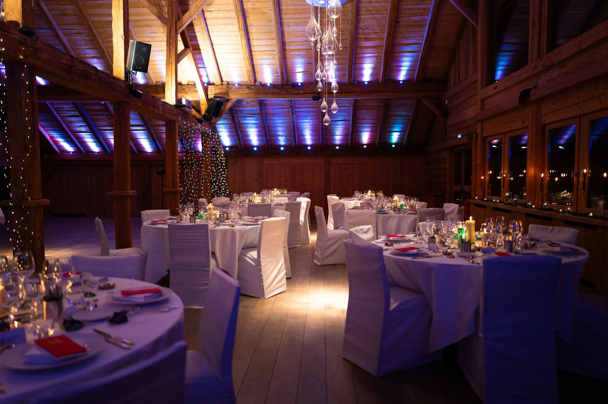 Salle de réception élégante avec tables rondes, nappes blanches, chaises avec housses blanches, décorations de bougies et lumières d'ambiance dans une pièce en bois avec plafond voûté.