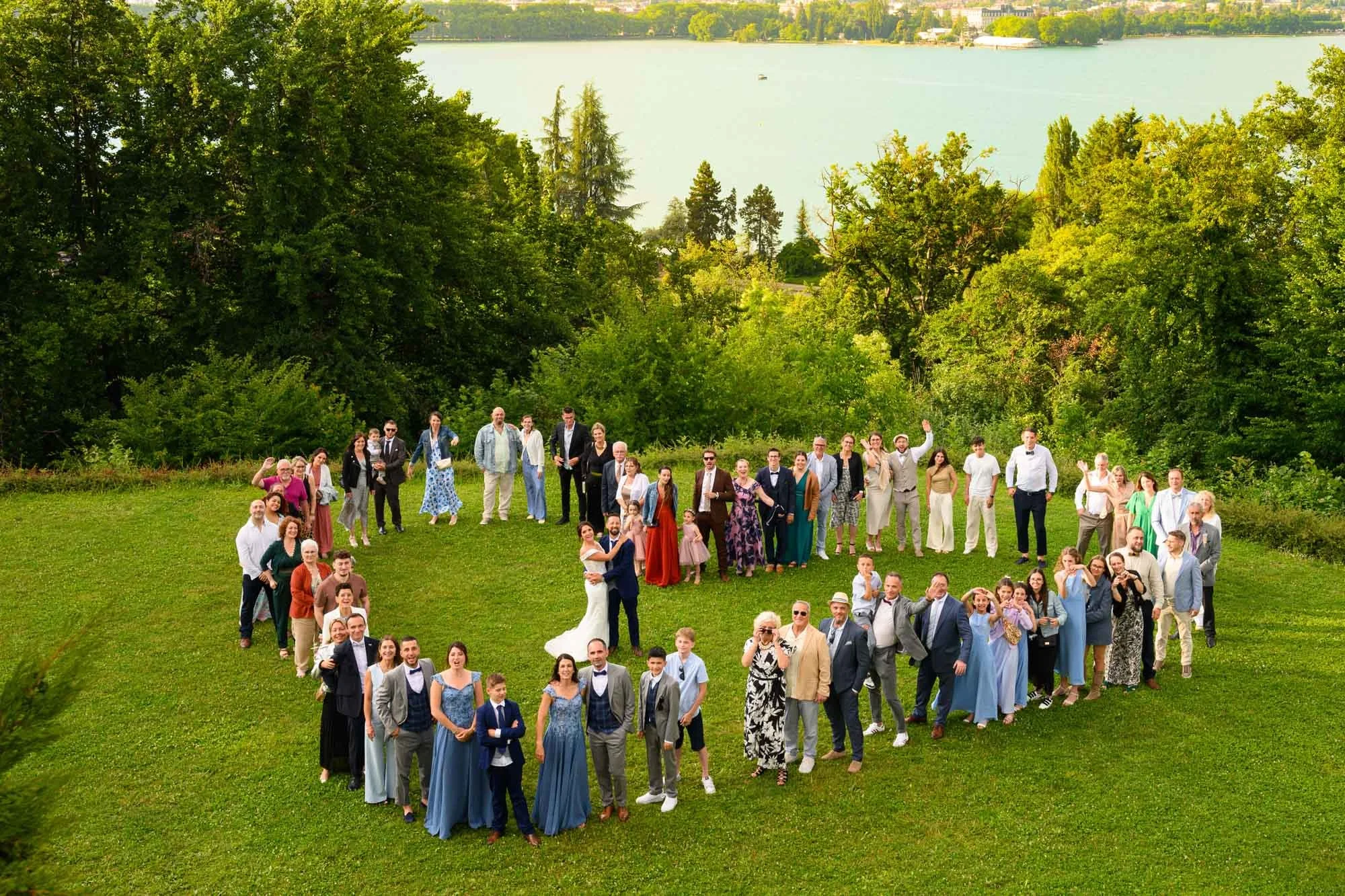 Groupe de personnes formant un cœur sur une pelouse près d'un lac avec des arbres et une ville en arrière-plan.