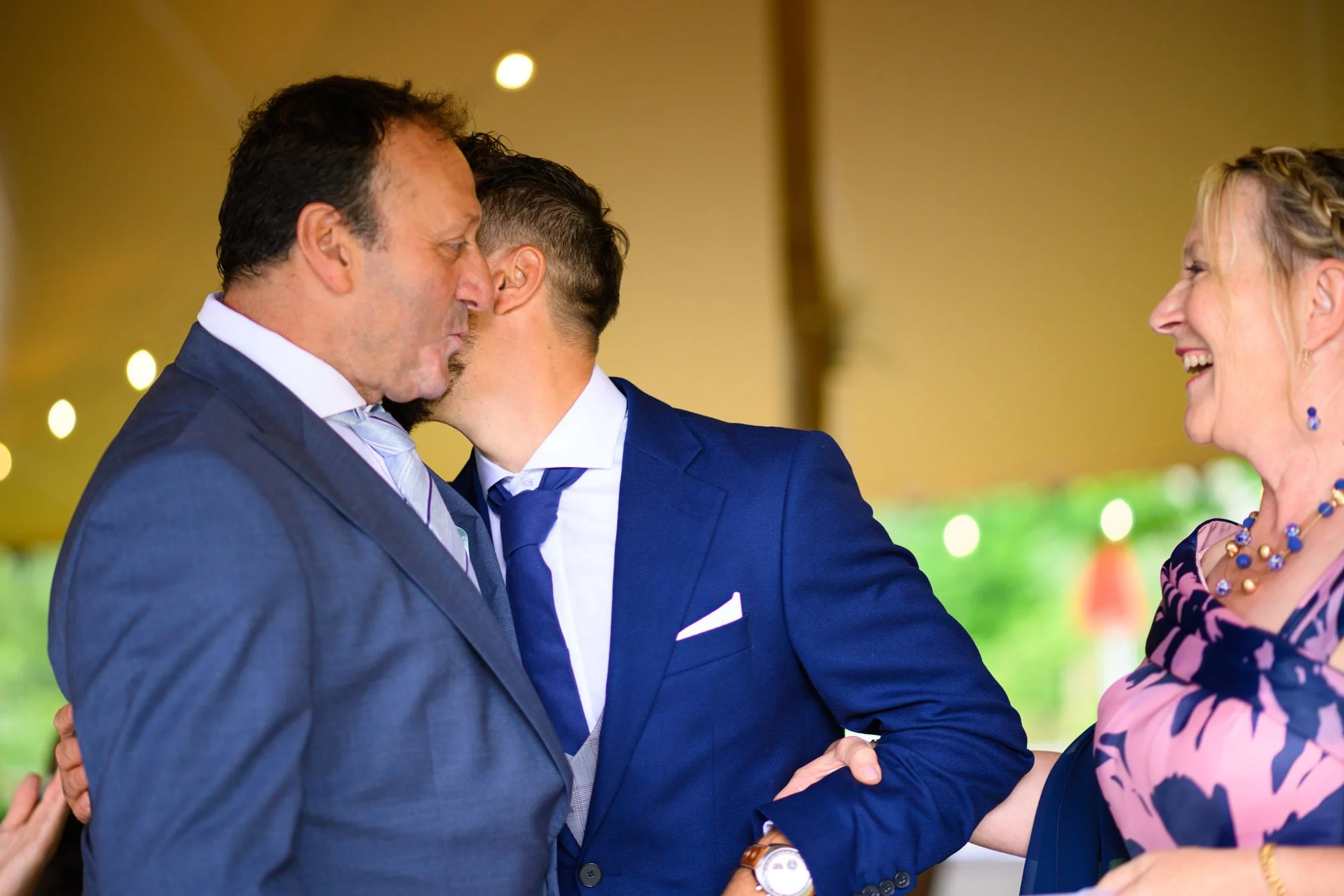 Trois personnes en costume se saluent et rient lors d'un événement. Deux hommes et une femme, dans un cadre festif. La femme porte un collier coloré.