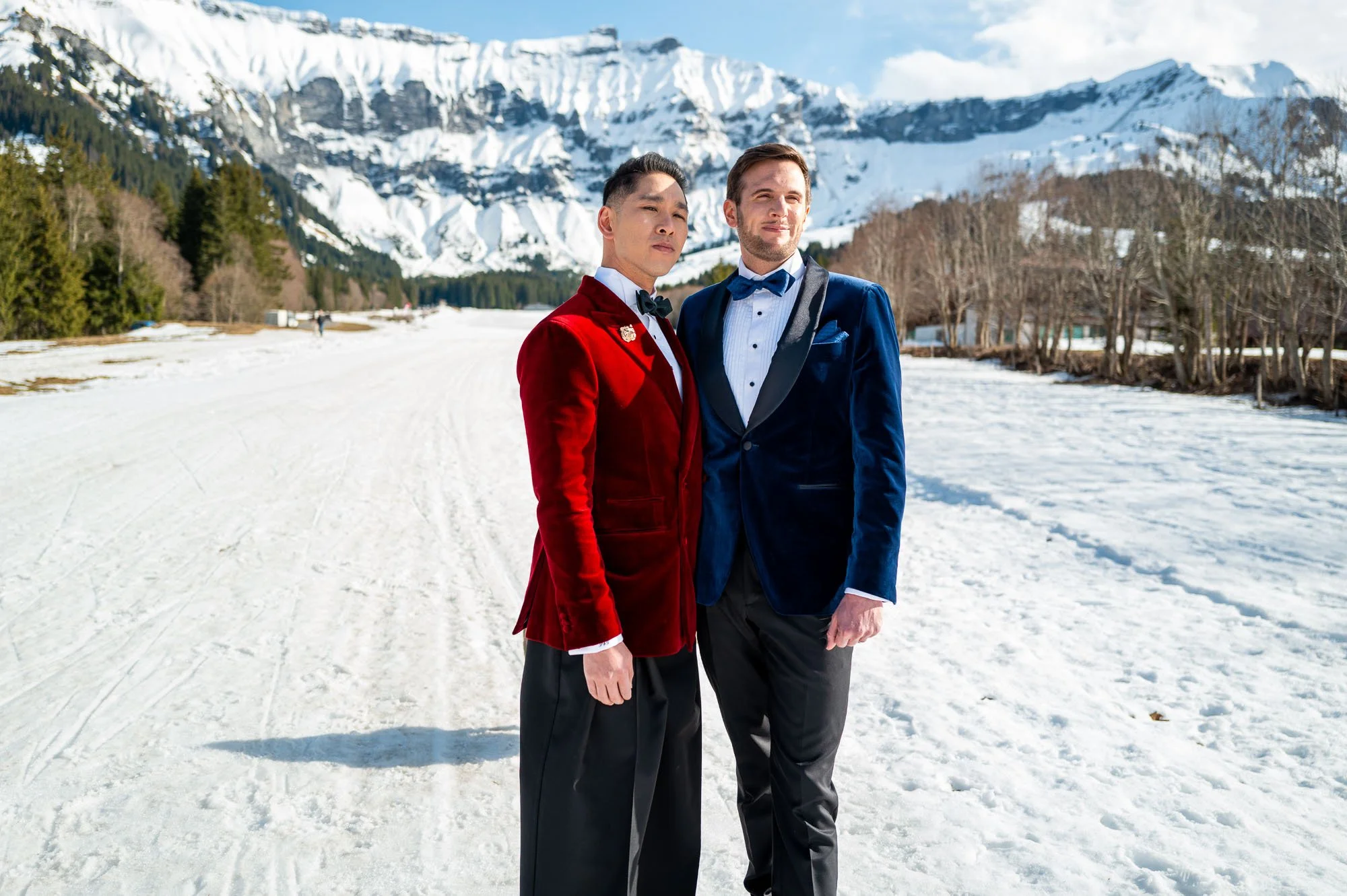 Deux hommes en costumes élégants posent dans la neige, avec des montagnes enneigées en arrière-plan.