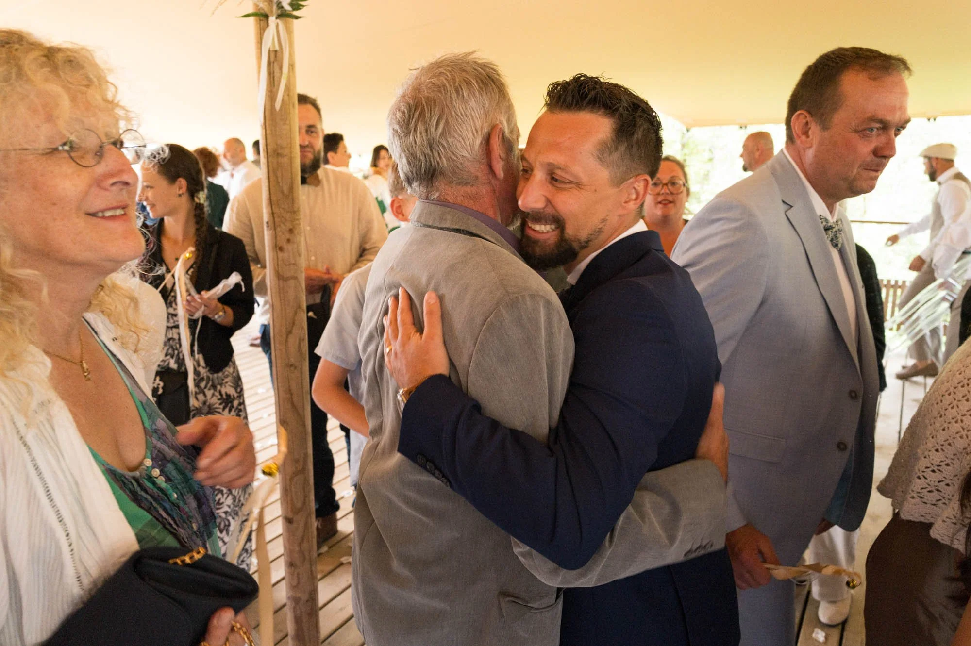 Deux hommes, l'un plus âgé, s'embrassent lors d'une célébration, entourés de plusieurs personnes souriantes dans un grand espace couvert.