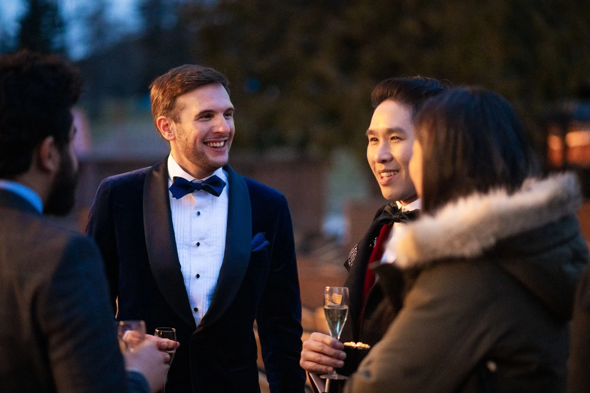 Groupe de quatre personnes habillées élégamment, discutant et riant lors d'une soirée en plein air, tenant des verres de champagne.