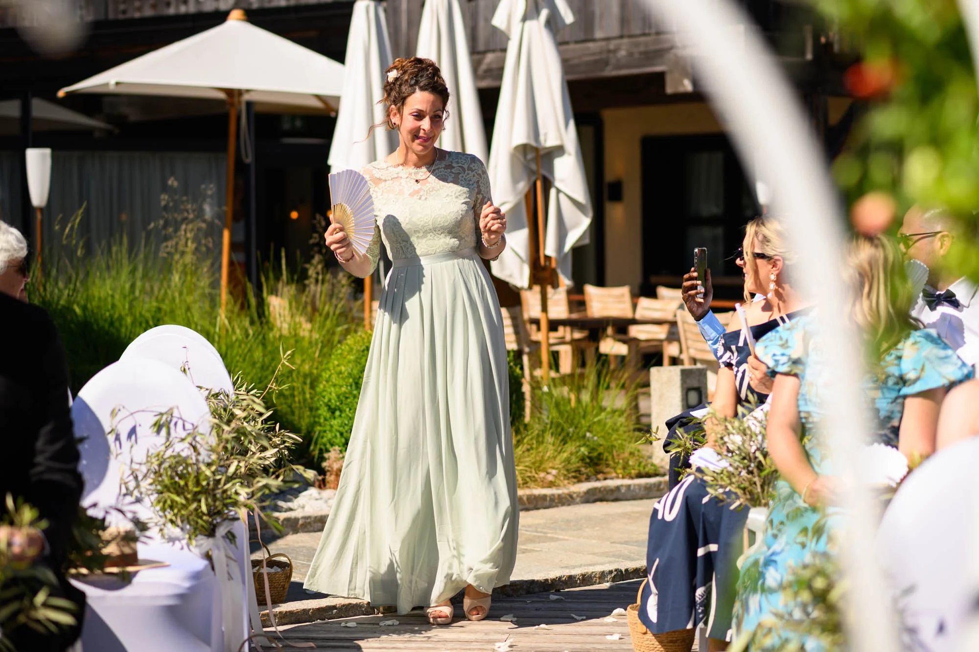 Femme élégante en robe longue à l'extérieur, tenant un éventail, entourée d'invités lors d'une cérémonie ou célébration en plein air sous un soleil brillant.