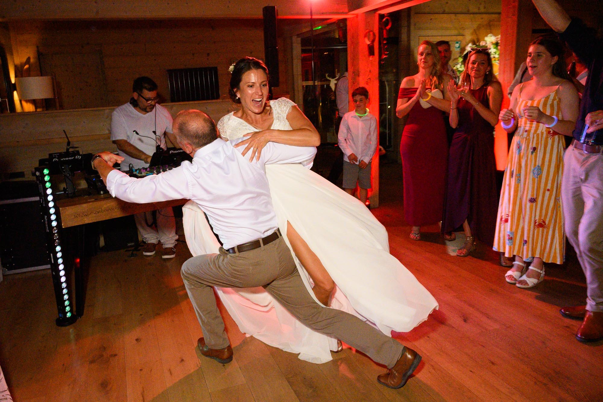 Une femme en robe de mariage danse joyeusement avec un homme lors d'une célébration, entourée d'invités qui applaudissent dans une ambiance chaleureuse et festive.