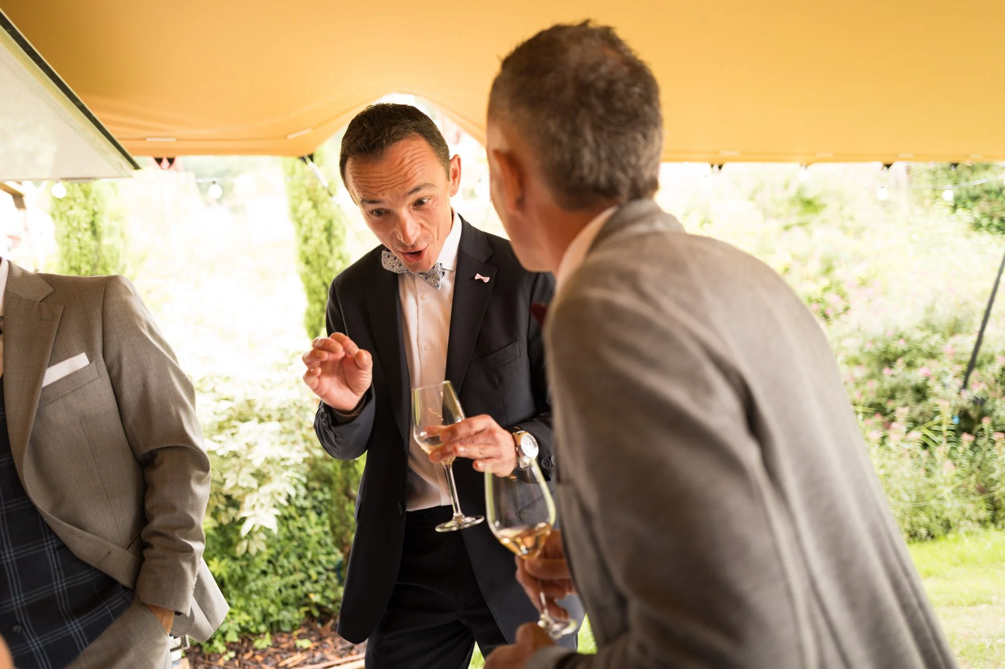 Trois hommes habillés en costume discutent lors d'une fête en extérieur, tenant des verres de champagne, avec un décor de jardin en arrière-plan.