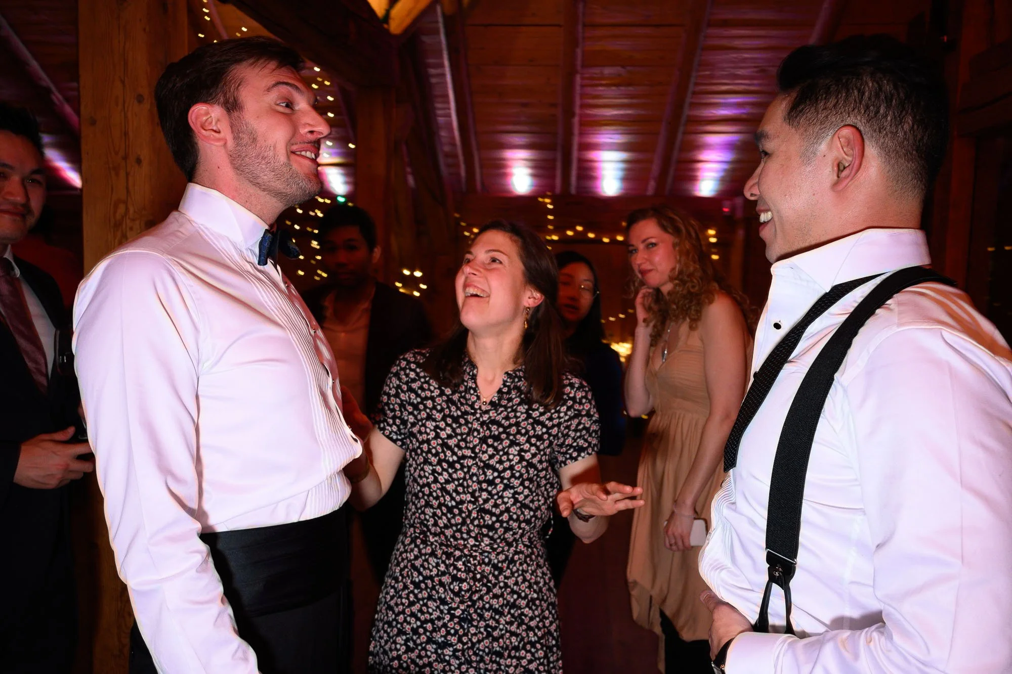 Groupe de personnes souriantes et discutant lors d'une fête ou d'un événement social, dans un décor chaud en bois avec des lumières scintillantes.