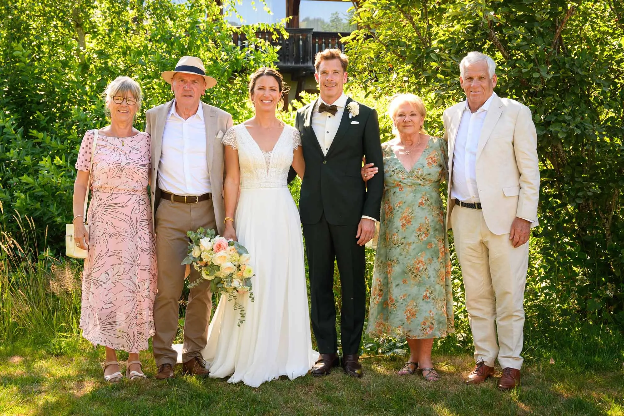 Groupe de six personnes posant pour une photo de mariage en plein air, avec un couple de mariés au centre, entourés de leurs proches, sous un arbre verdoyant.