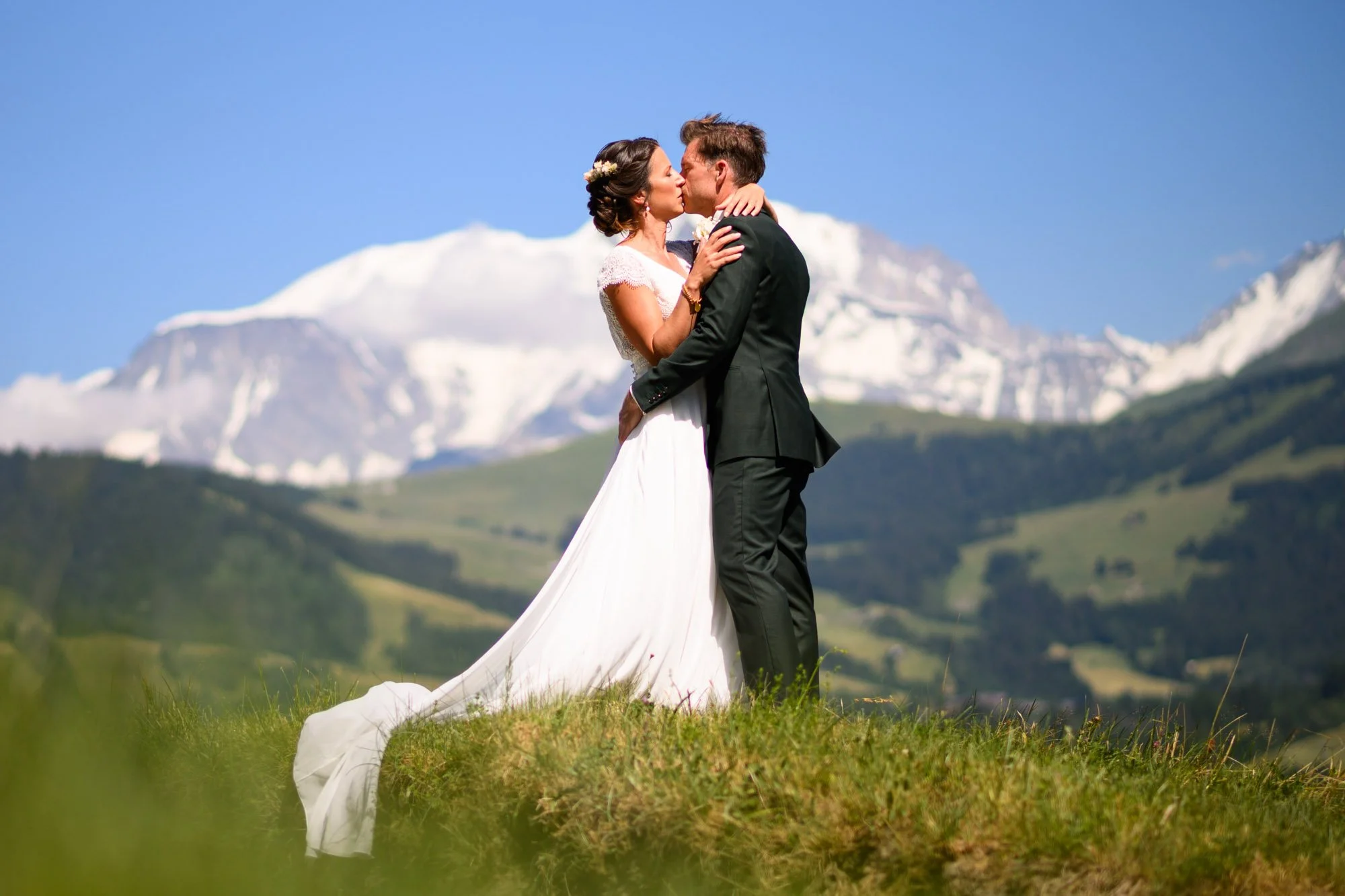 Un couple de mariés s'embrassant dans un paysage montagneux, avec des montagnes enneigées en arrière-plan et un ciel bleu clair.