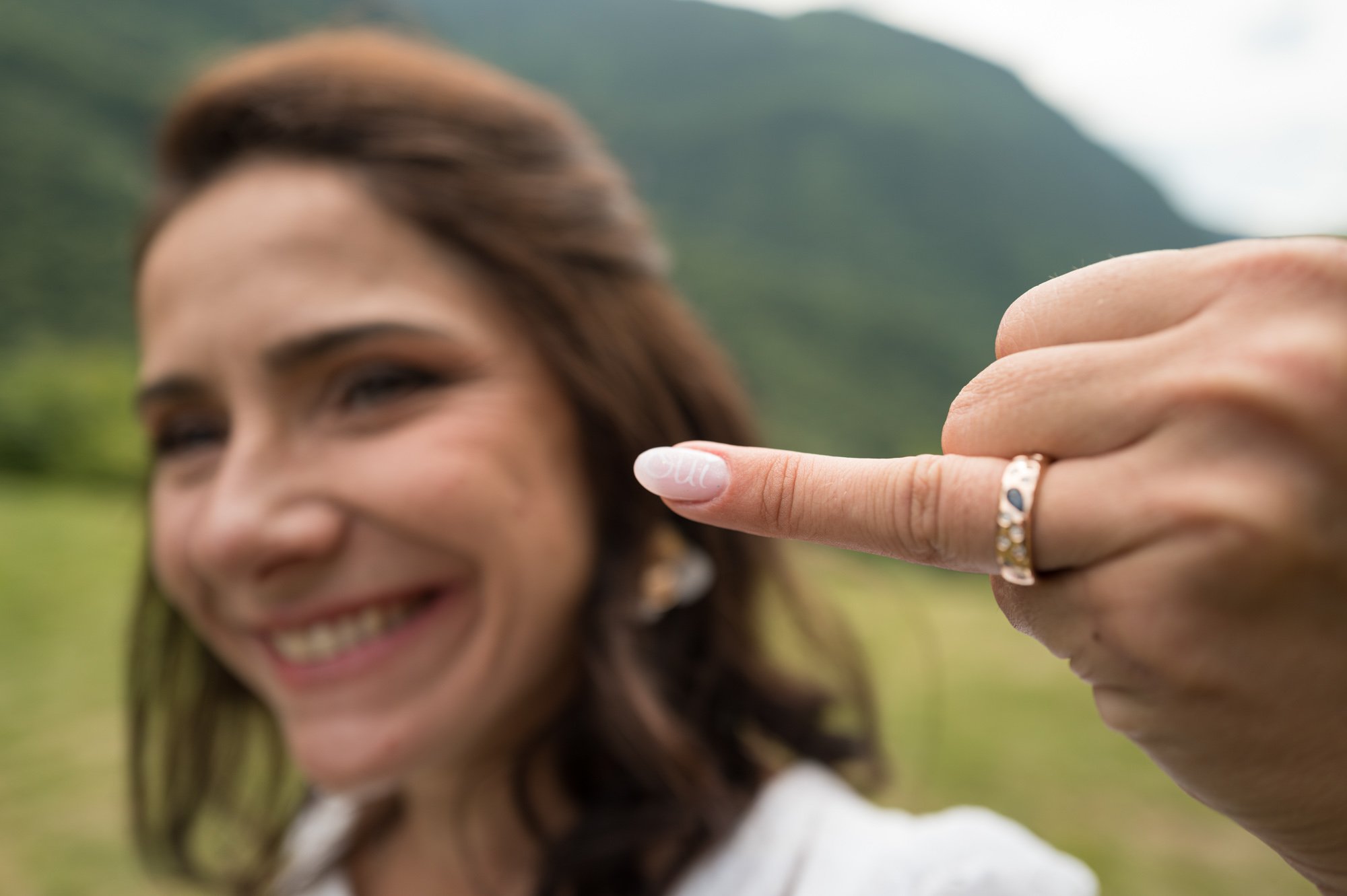 Jeune femme souriante tenant sa main avec un vernis à ongles transparent et un anneau en or, avec un paysage naturel en arrière-plan.
