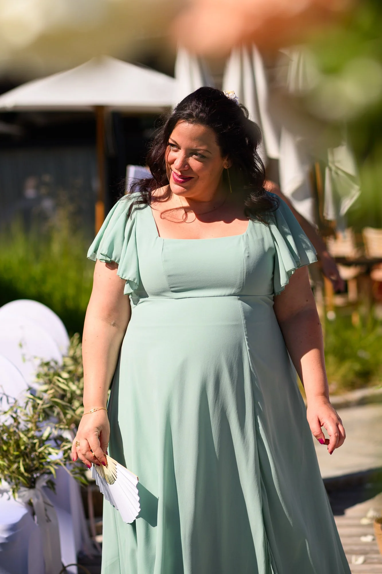 Une femme portant une robe vert pâle, souriant, lors d'une occasion extérieure ensoleillée.