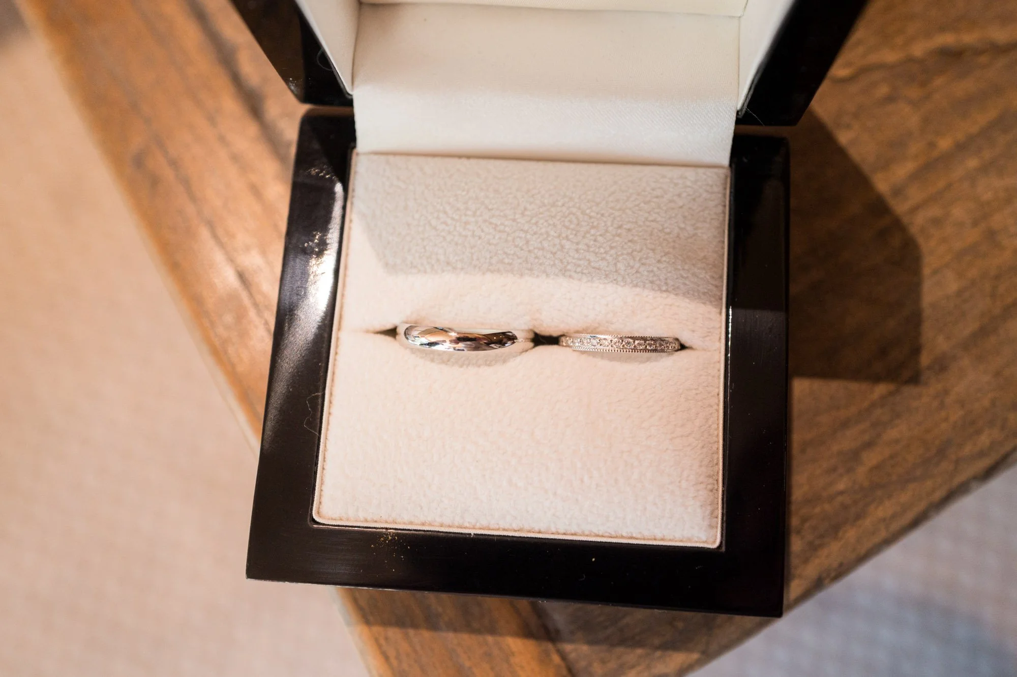 Deux alliances dans une boîte à bijoux noire avec intérieur en velours beige, posée sur une surface en bois.