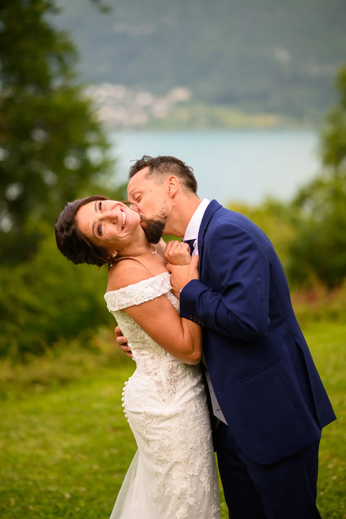 Un couple lors d'un mariage, avec la mariée en robe blanche et le marié en costume bleu, partageant un moment d'intimité dans la nature, avec un lac et des montagnes en arrière-plan.