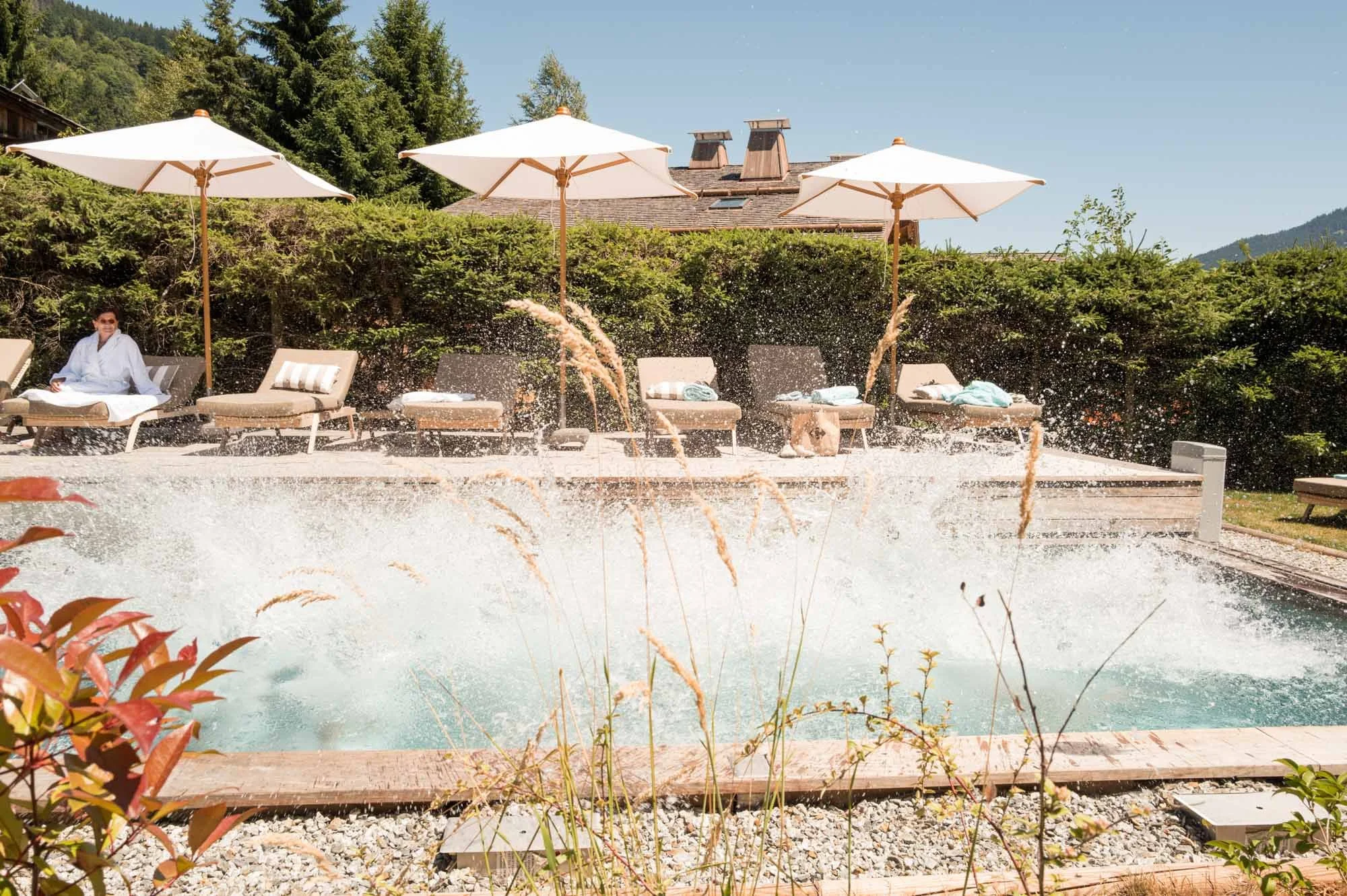 Piscine avec deux jets d'eau, parasols blancs, chaises longues avec serviettes, un homme en peignoir, végétation dense en arrière-plan.