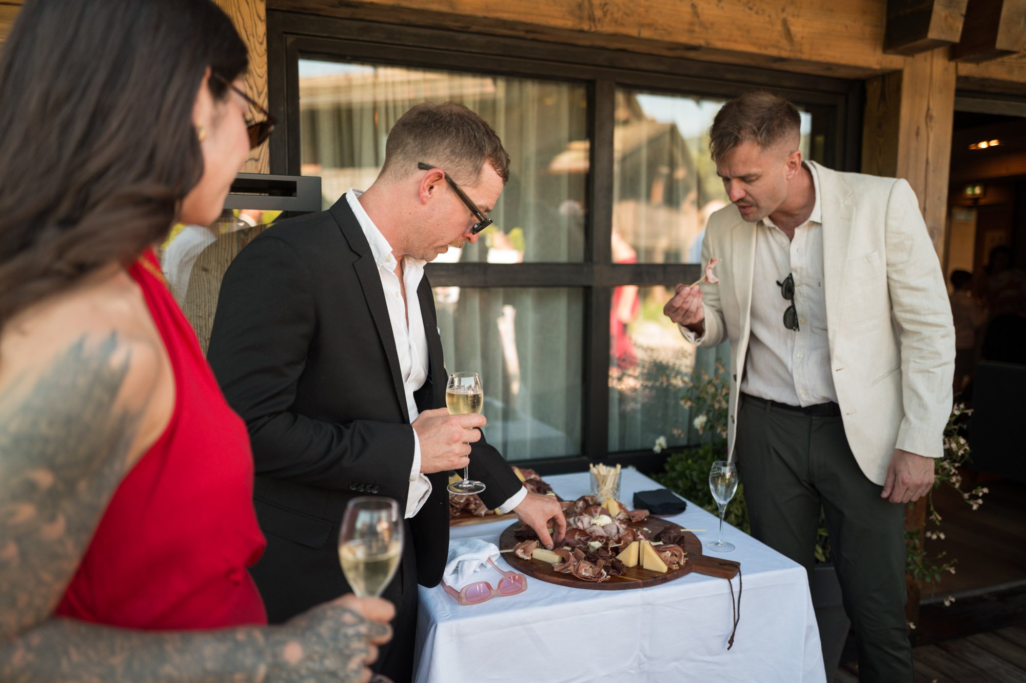 Groupe de trois personnes dégustant un plateau de charcuterie avec du fromage et des verres de vin blanc lors d'une fête ou événement social à l'extérieur.