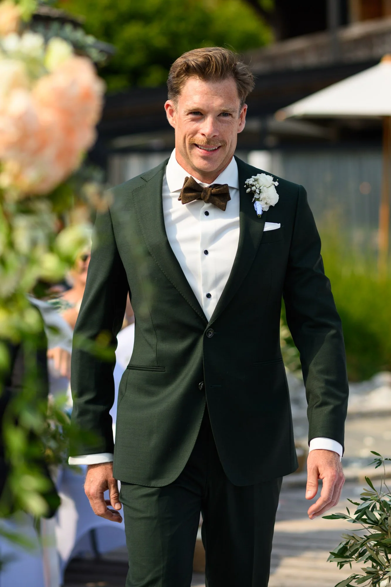 Un homme en costume de mariage noir avec une chemise blanche, un nœud papillon marron et une boutonnière blanche, souriant lors d'une cérémonie en plein air.