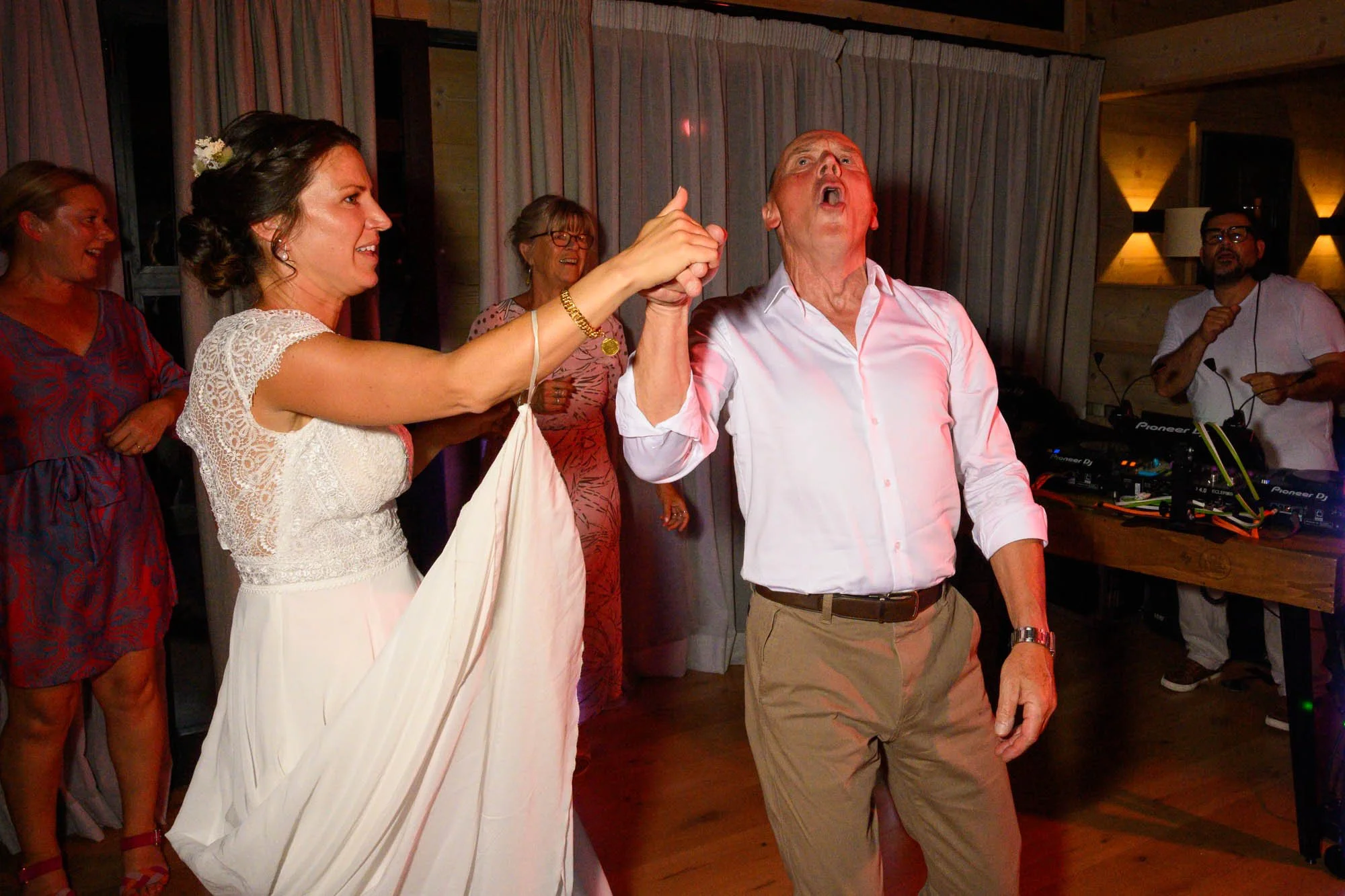 Une femme en robe de mariée dance avec un homme lors d'une fête, d'autres personnes regardent et sourient en arrière-plan.