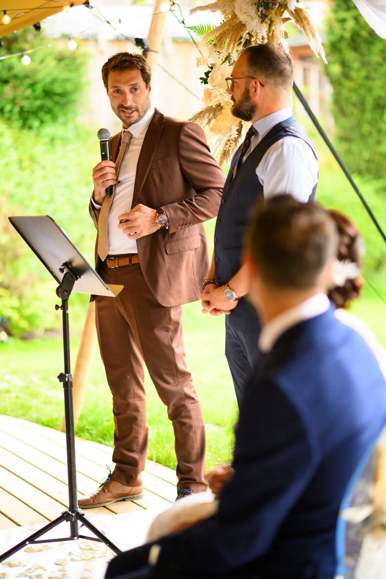 Un homme en costume marron parle lors d'une cérémonie en extérieur, probablement un mariage, tandis qu'un autre homme en costume sombre lui tient la main et écoute. Il y a une femme assise en arrière-plan, vue de dos. La scène est décorée de fleurs e