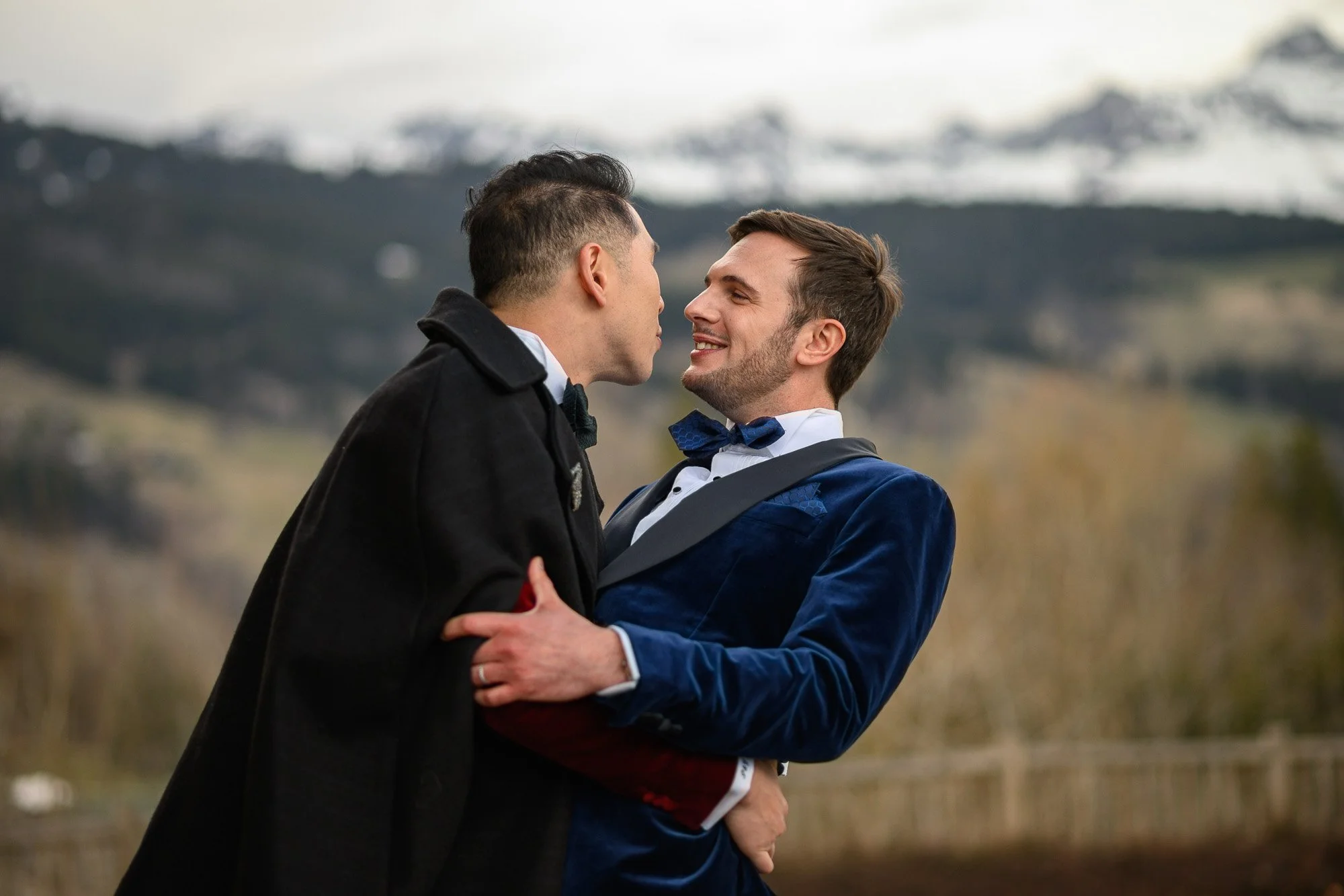 Deux hommes habillés en costumes élégants se tiennent proches, souriants, avec un arrière-plan de montagnes et de nature.