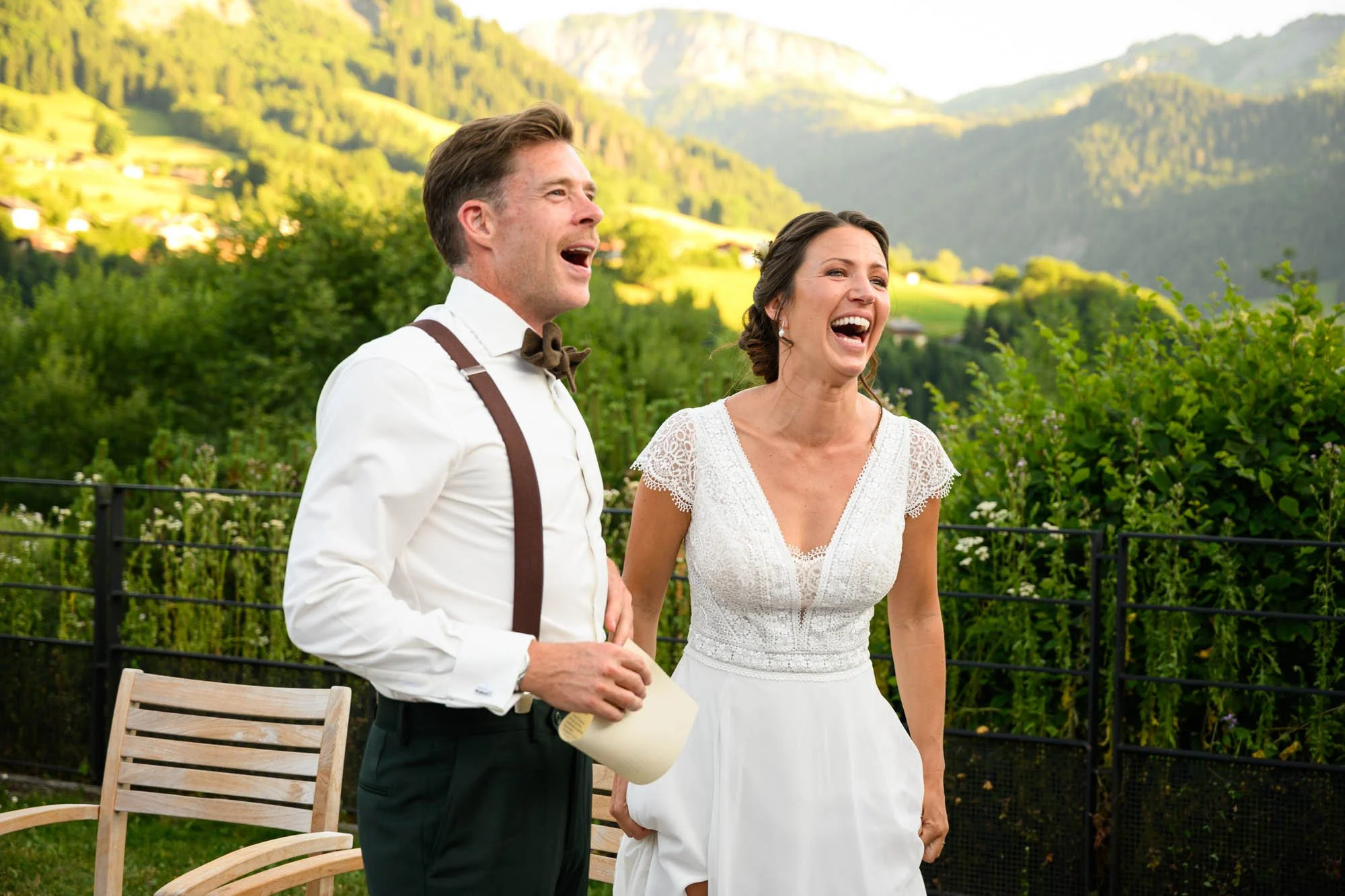 Un homme et une femme habillés pour un mariage, souriants et riants, à l'extérieur avec un paysage de montagnes et de verdure en arrière-plan.