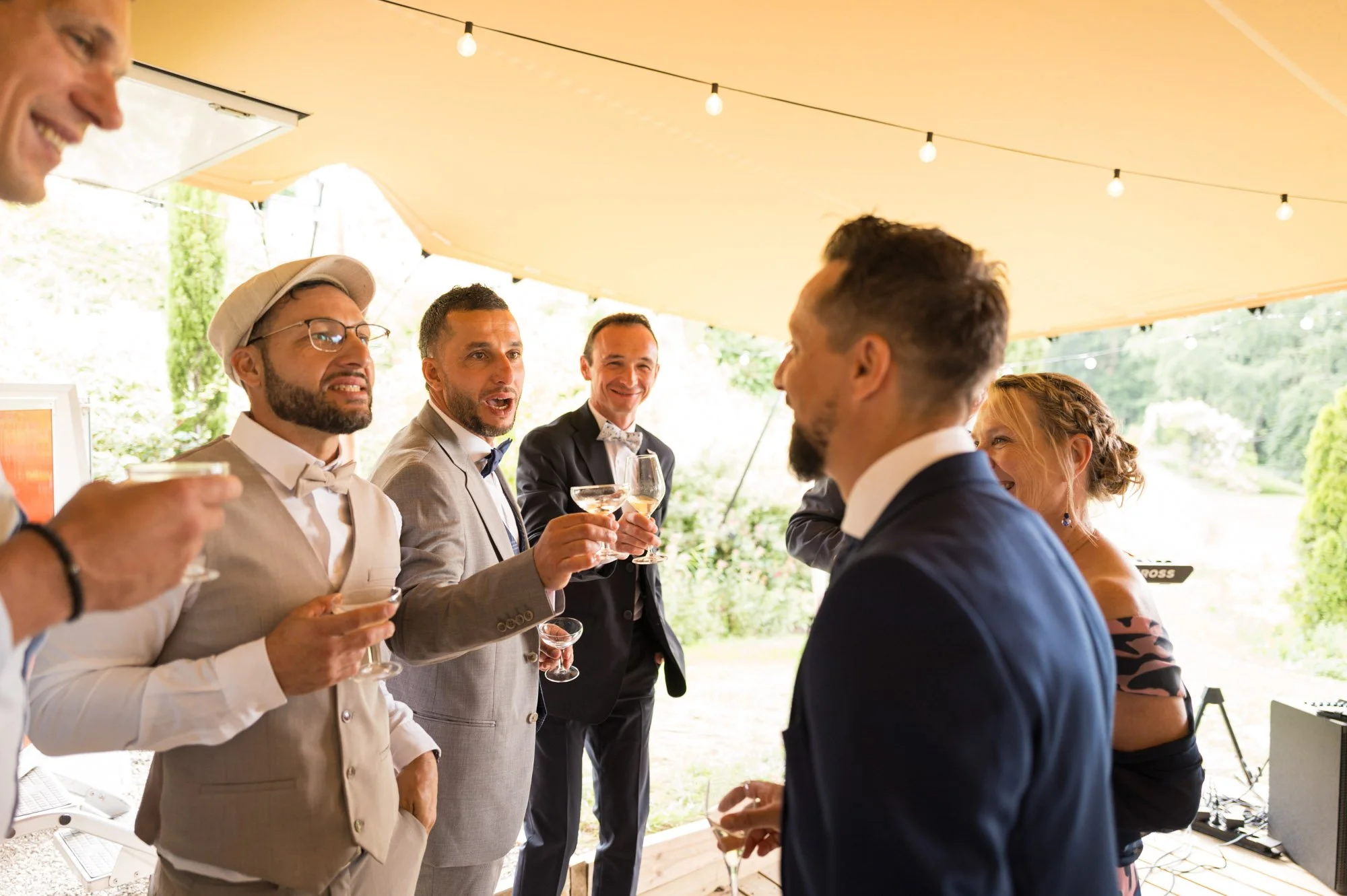Groupe de personnes habillées en costumes élégants, socialisant et dégustant des cocktails lors d'une célébration en plein air sous une tente avec des guirlandes lumineuses.