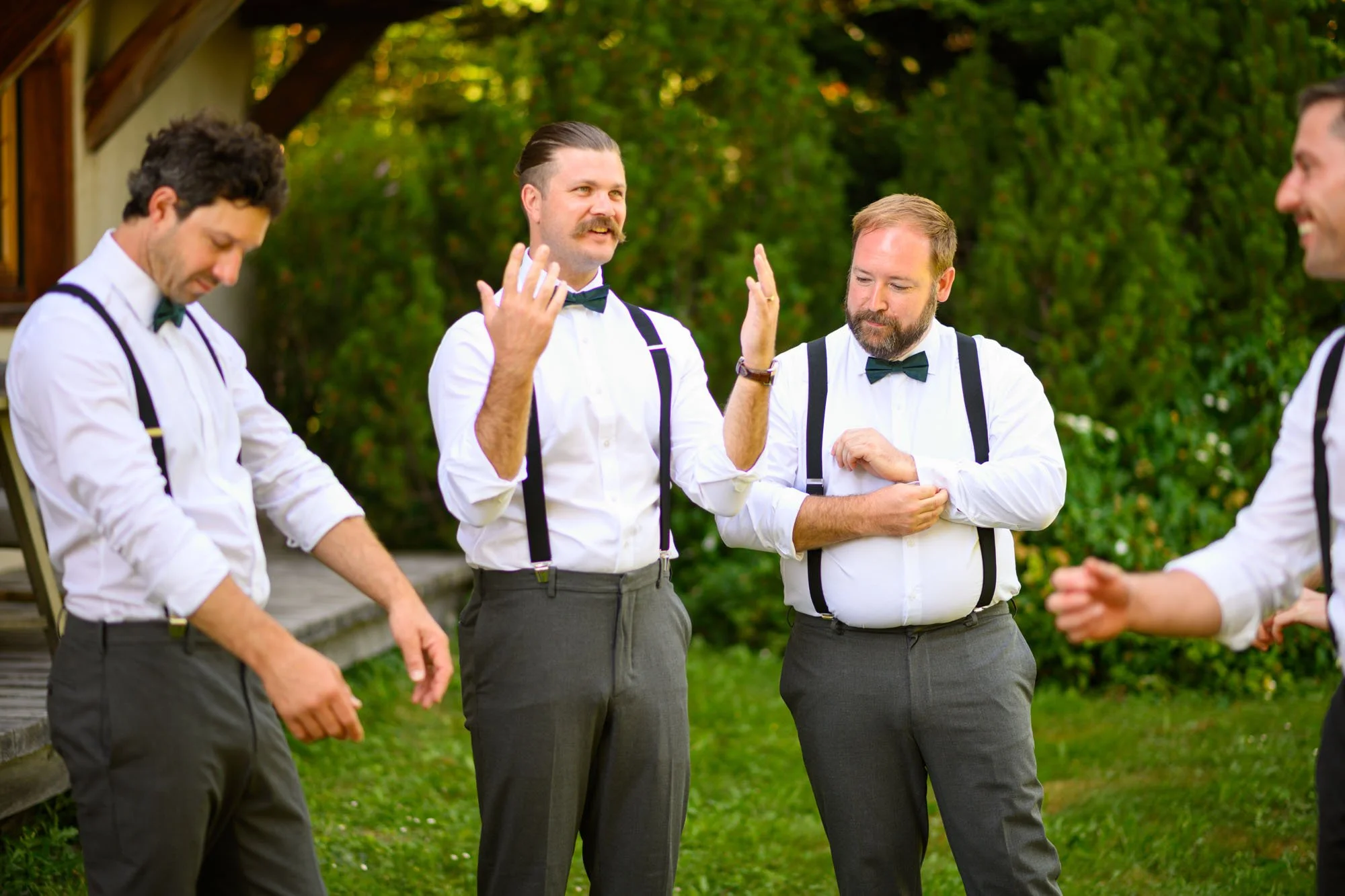 Quatre hommes habillés en tenue formelle avec chemises blanches, nœuds papillon et bretelles discutant lors d'un événement en extérieur, semble-t-il un mariage, avec un fond de végétation verte.