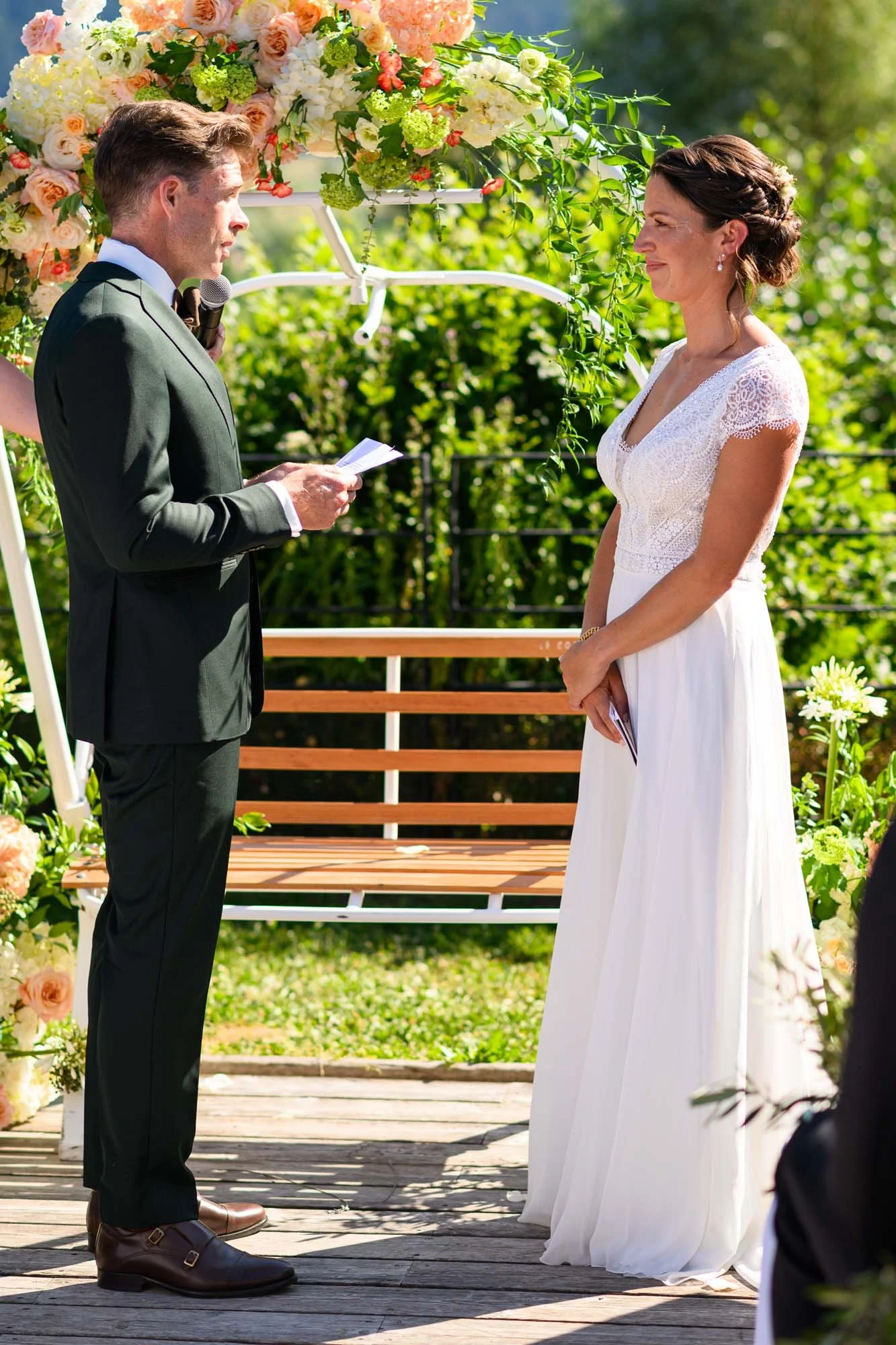Un couple en train de se marier lors d'une cérémonie en plein air, sous un décor floral, avec l'homme tenant un micro et lisant ses vœux, et la femme en robe blanche écoutant attentivement.