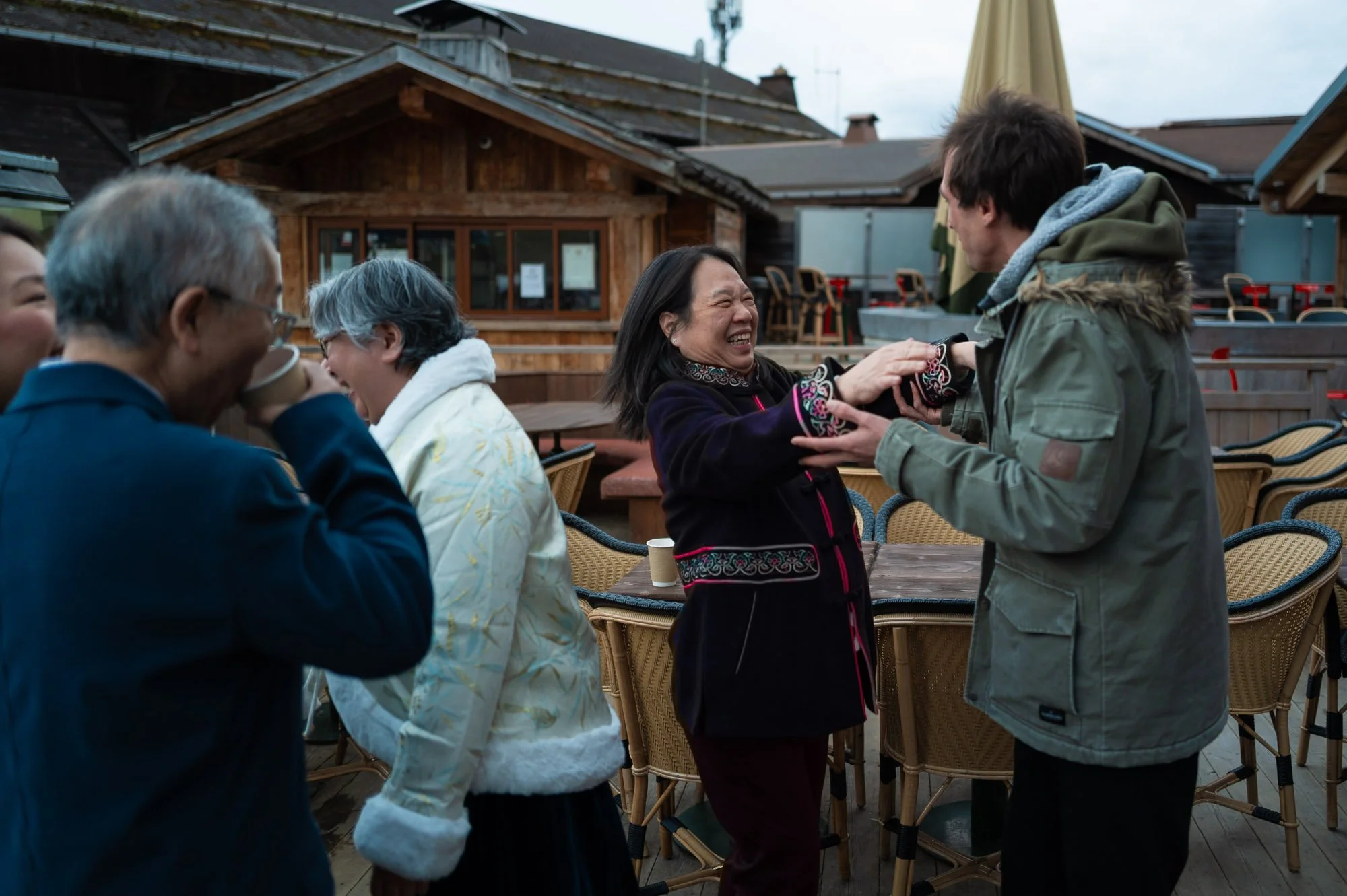 Groupe de personnes qui rient et partagent un moment joyeux sur une terrasse en plein air, avec des bâtiments en bois en arrière-plan.